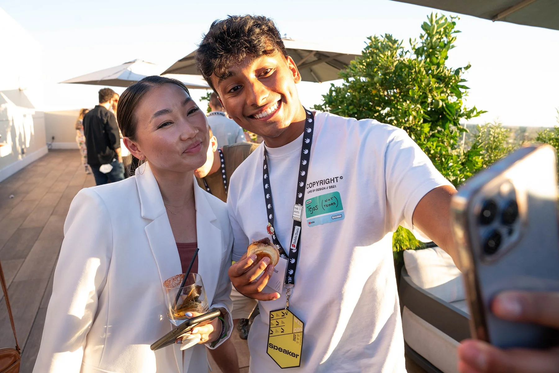 Two people taking a selfie at an outdoor event during sunset, with the woman holding a drink and the man holding a pastry, both smiling.