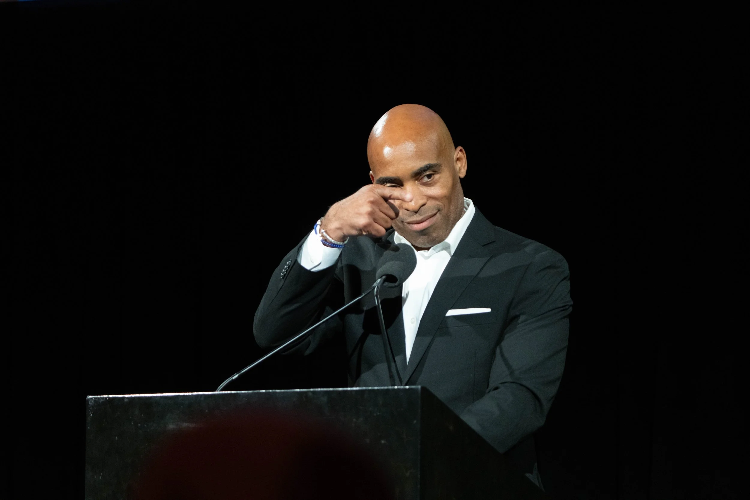 Man in a black suit and white shirt standing at a podium with a microphone, touching his eye, against a black background.