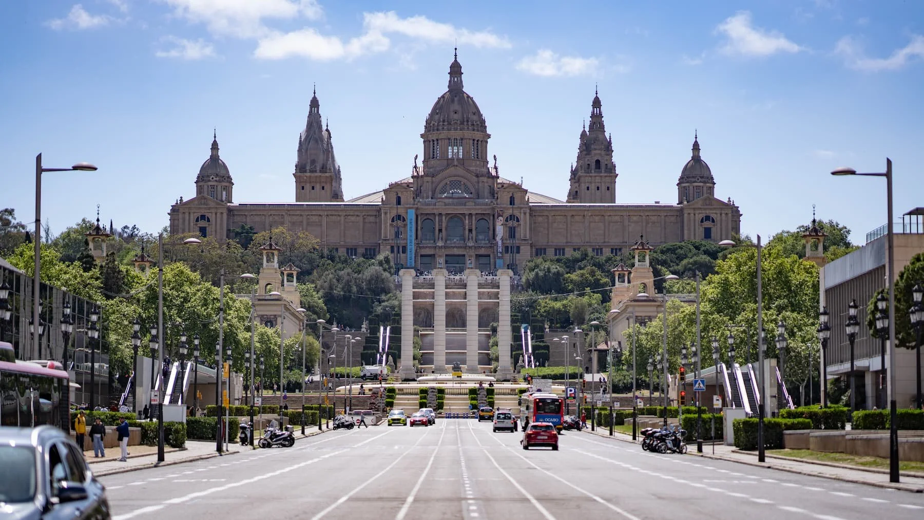 Wide view down a broad avenue in Barcelona toward the domed Palau Nacional on Montjuïc, home of the MNAC museum, under a blue sky with light traffic on the road.