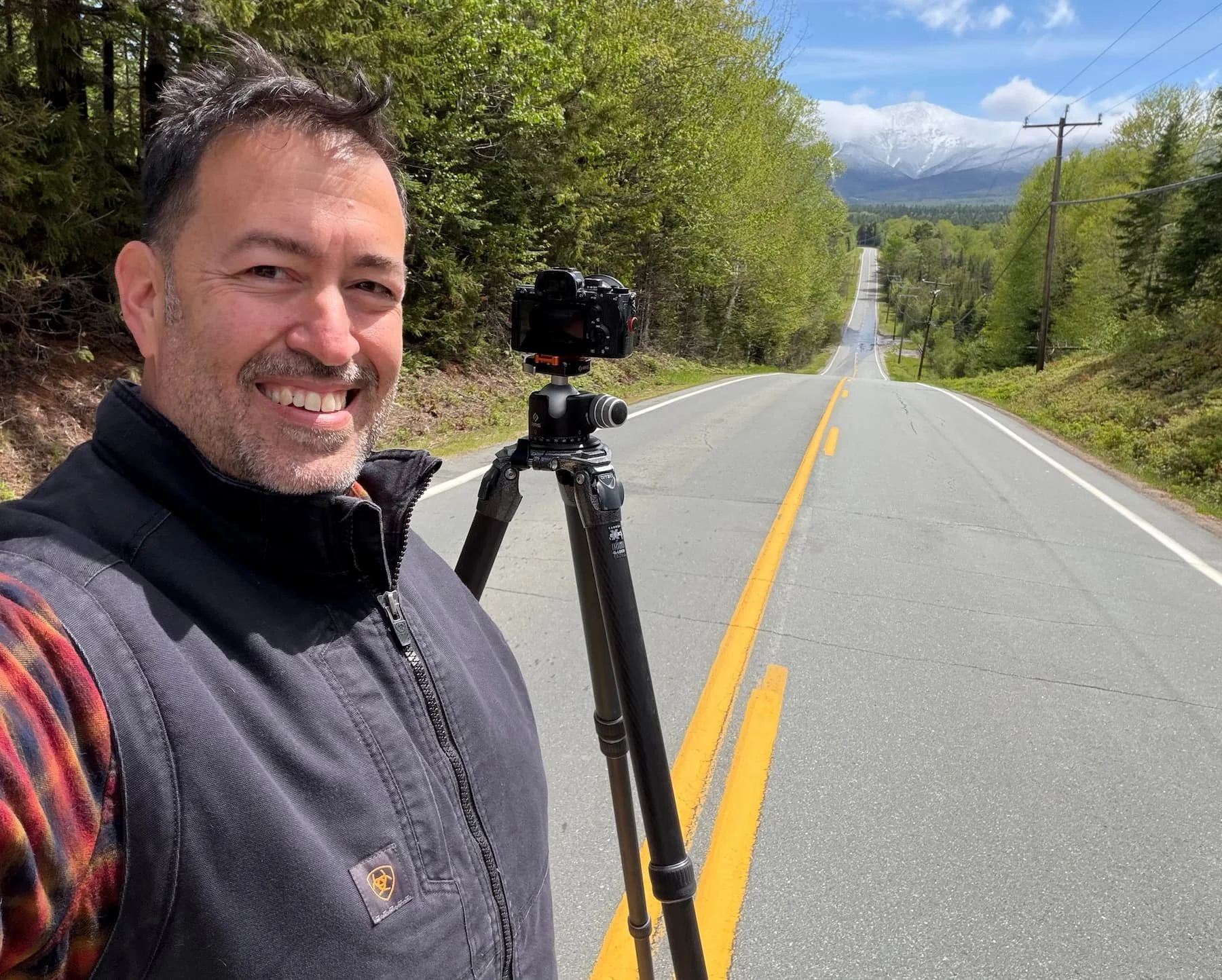 Anthony Quintano smiling and taking a selfie on a New Hampshire road, camera and tripod set up beside him, with a snow-capped Mount Washington in the distance on the first day of summer 2026.