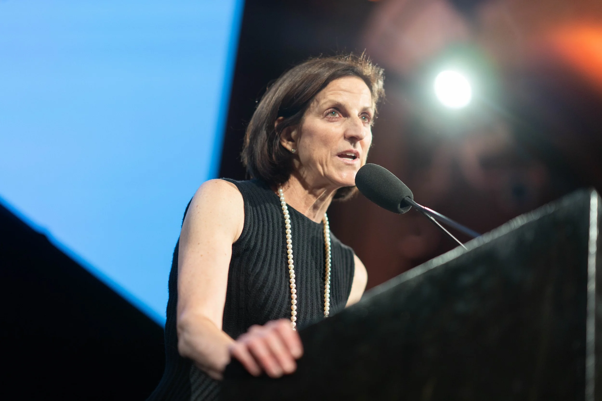 Woman with dark hair wearing a black sleeveless dress and a pearl necklace speaking at a podium with a microphone.
