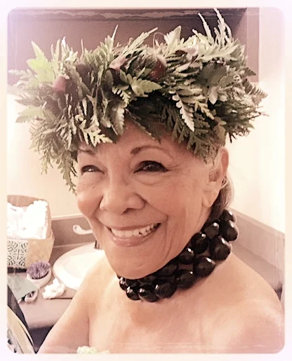 A smiling elderly woman wearing a leafy crown, a dark beaded necklace, and minimal clothing, possibly at a celebration or cultural event.