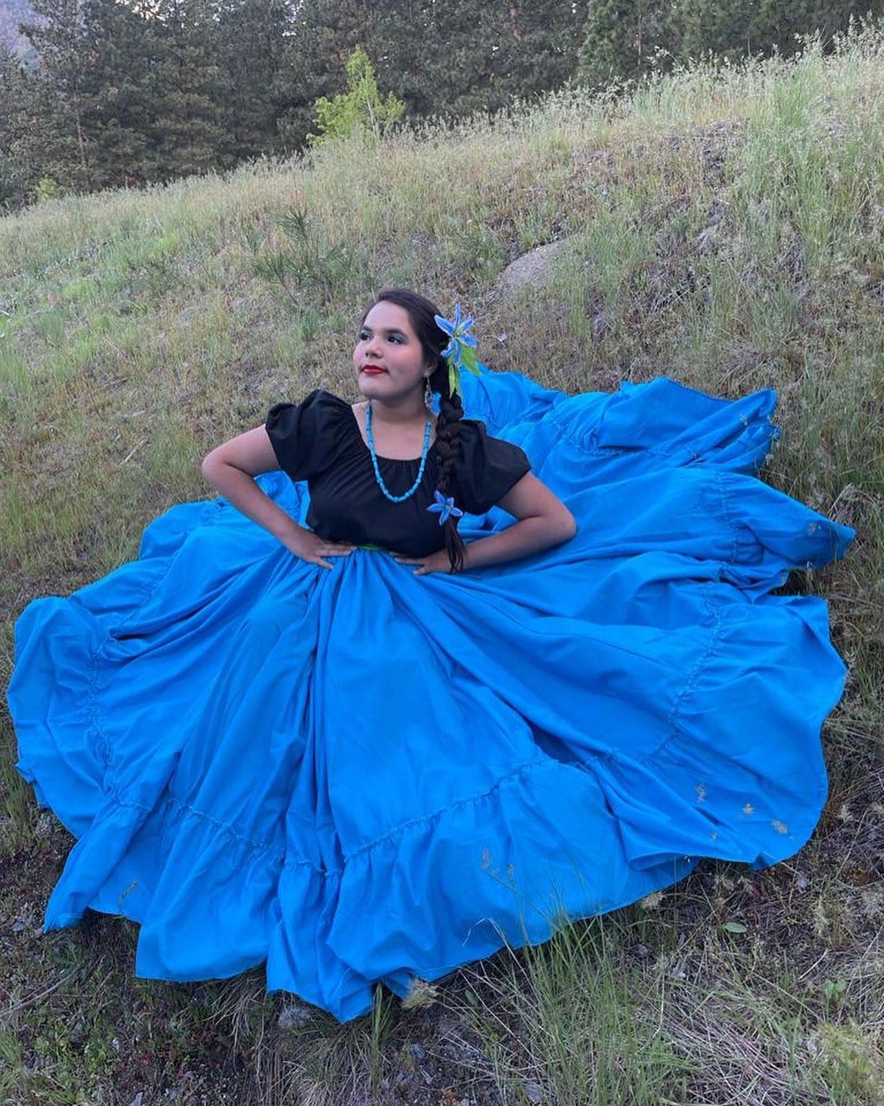 A woman lying on the grass wearing a black top and a bright blue skirt with a flower in her hair and necklace, outdoors with trees in the background.