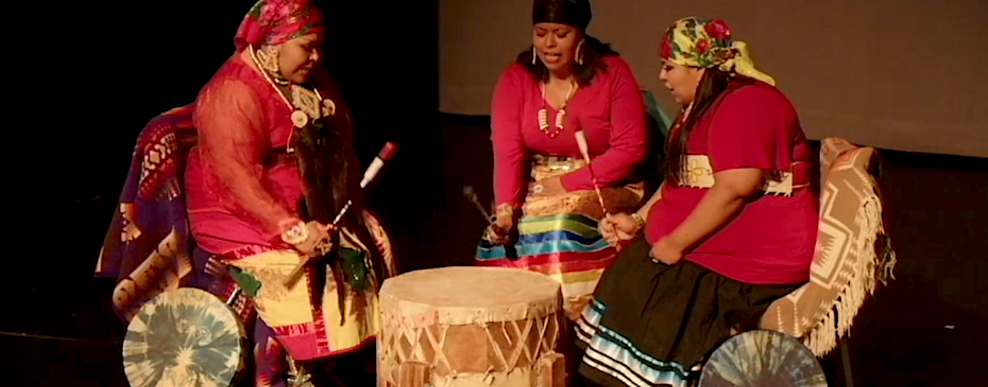 Three women sitting around a drum, dressed in colorful traditional African attire, participating in a cultural activity.