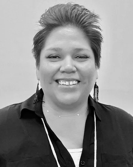 A woman with short hair smiling, wearing earrings, a dark top, and a necklace.  Mary Big Bull Lewis 