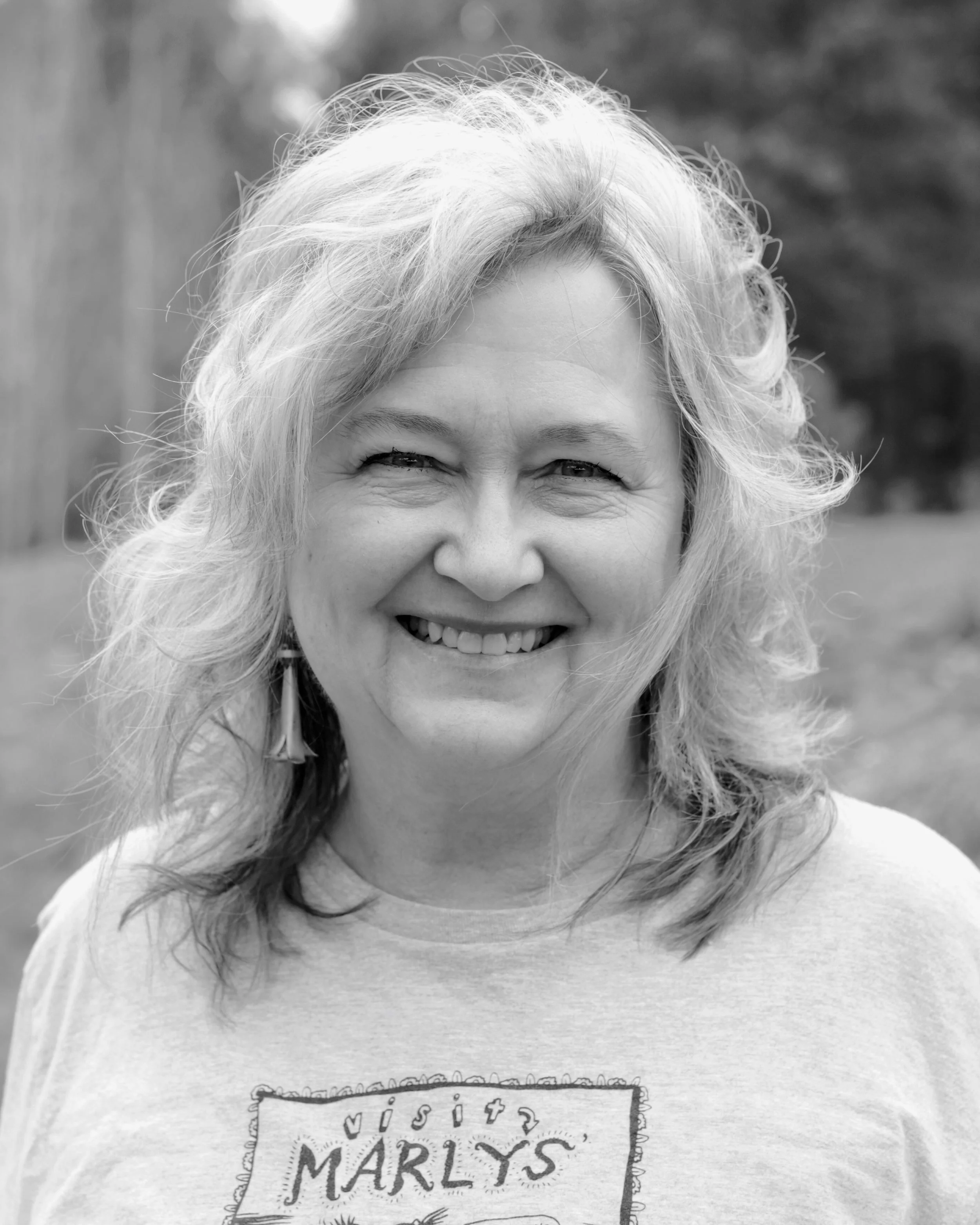 Black and white photo of a smiling woman with curly hair, wearing a t-shirt with the word "Marlys" on it, outdoors with blurred background.  Marile Sexson Kunkel 