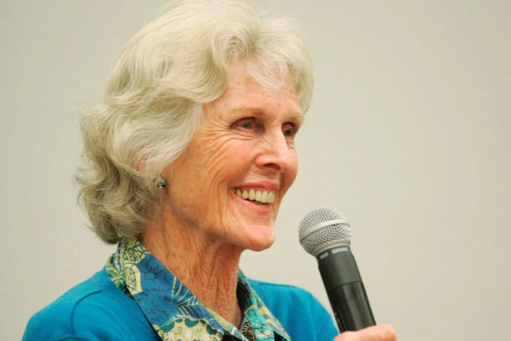 An elderly woman with white, curly hair speaking into a microphone, smiling, wearing a blue top and patterned shirt, standing against a plain beige wall.  Harriet Bullit,  Harriet Stimson Bullit, Creative person, Icicle Creek Center for the Arts  
