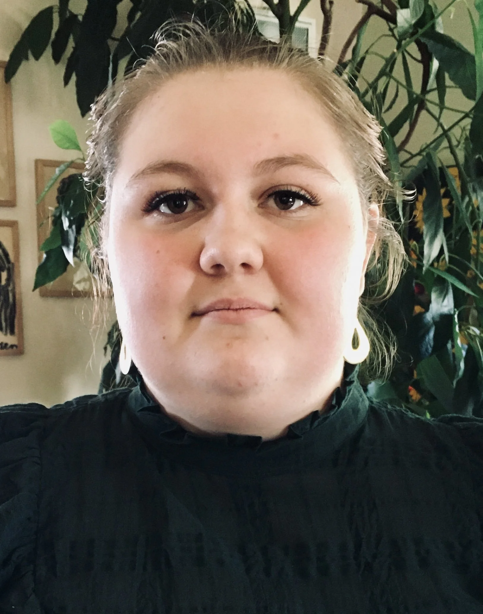 Close-up of a young woman with blonde hair and hoop earrings, wearing black clothing, standing in front of green leafy plants indoors.