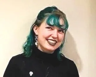 A woman with dark hair and teal highlights smiling, wearing a black top and jewelry, against a plain background.  Hannah Blank, Tech. 