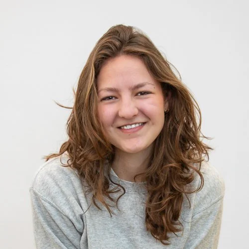 een jonge vrouw met bruin, golvend haar die lacht, gedragen in een grijze trui, op een witte achtergrond