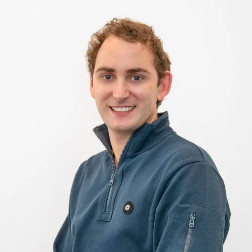 Portret van een jonge man met lichtbruine krulletjes, een blauwe jas en een vriendelijke glimlach, staand voor een witte achtergrond.