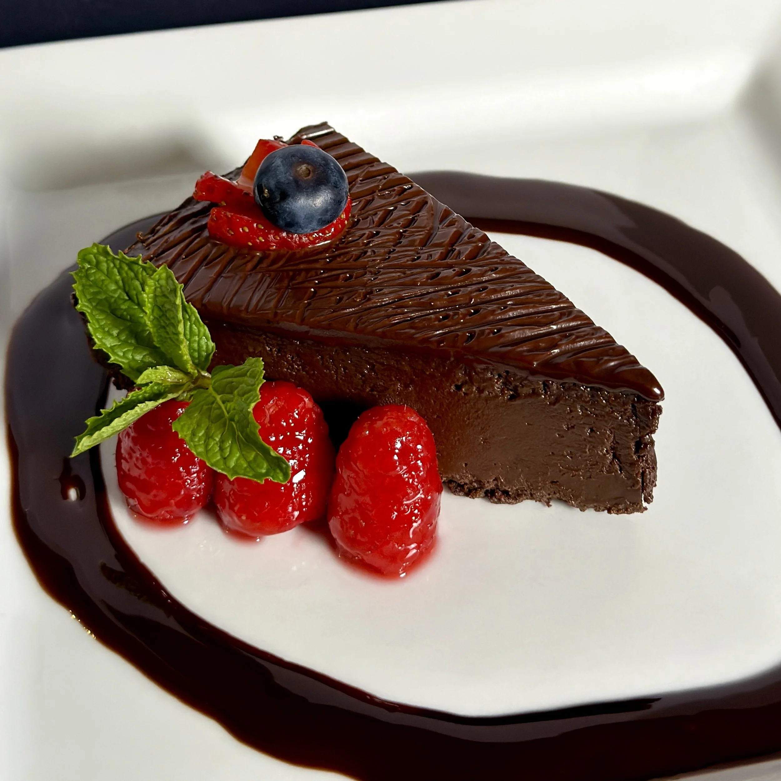 A slice of our flourless chocolate cake with glazed raspberries, blueberry, mint leaves, and chef's signaturechocolate drizzle. A popular dessert menu item.