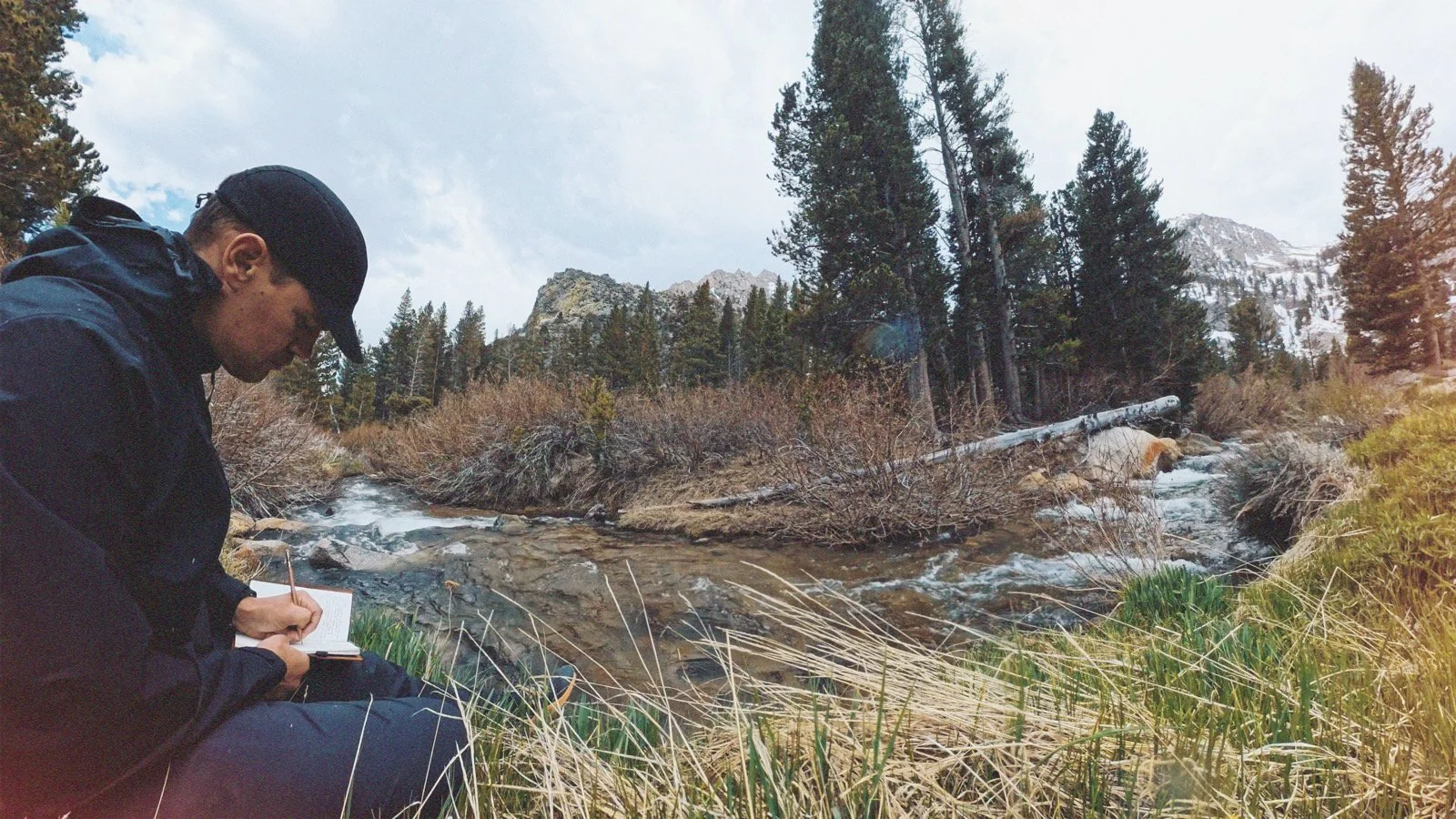 A person sitting outdoors near a stream writing in a notebook with mountains and trees in the background.