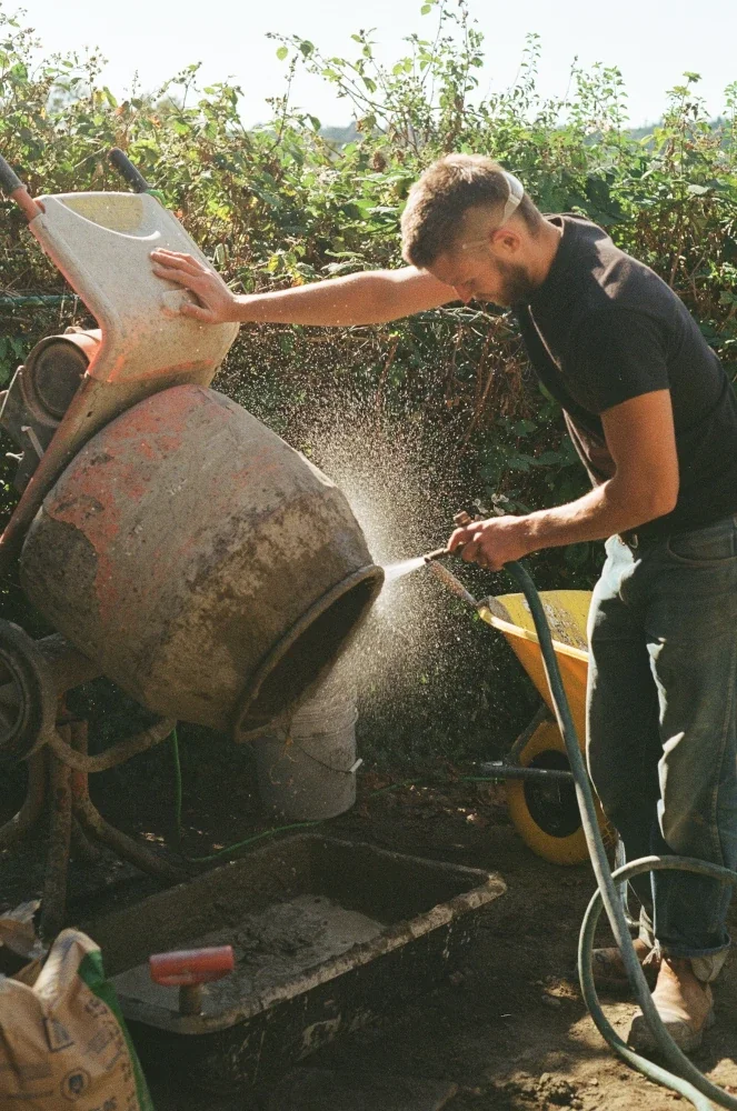 Stonemason Sandy hosing down a cement mixer, surrounded by greenery