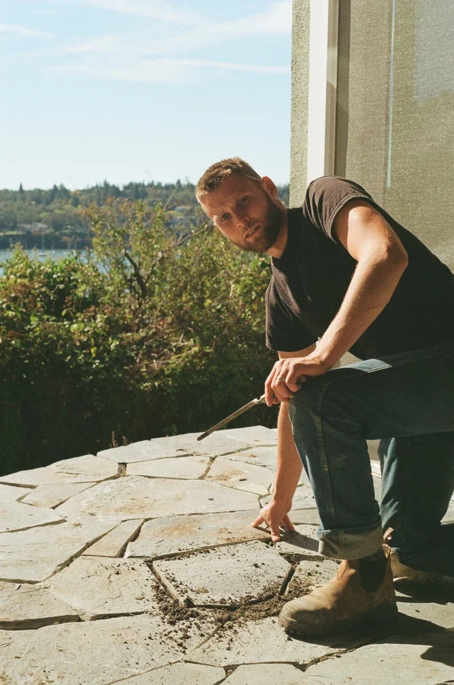 Stonemason Sandy kneeling over a stone patio with a landscape view of Victoria in the background
