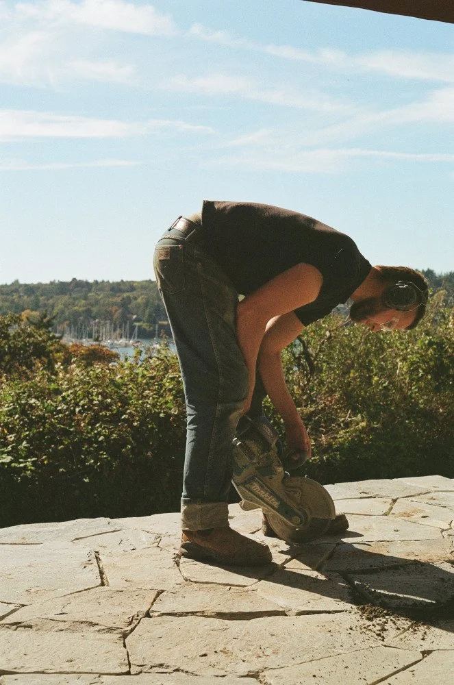 Stonemason Sandy leaning over a stone patio with a powersaw with a landscape view of Victoria in the background