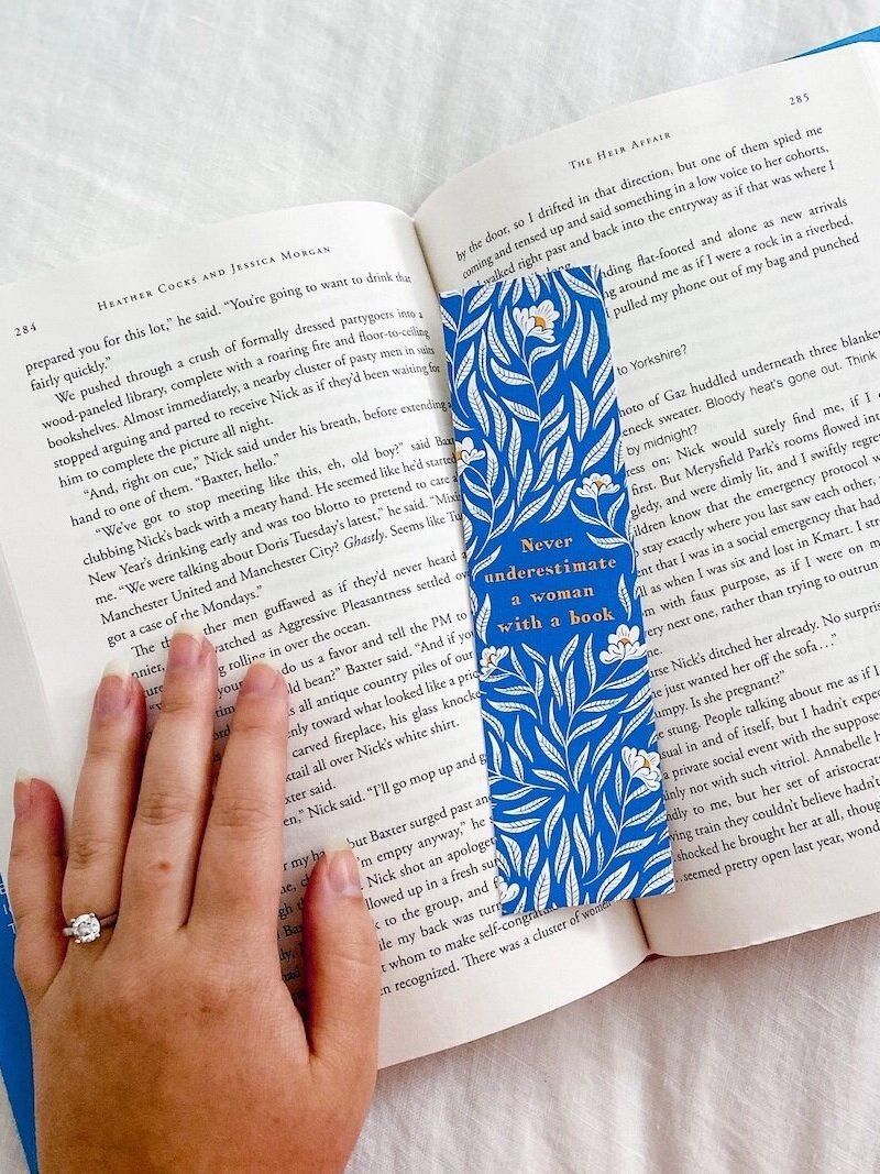 A book with a blue bookmark being held open by Phoebe's hand, an engagement ring on her ring finger