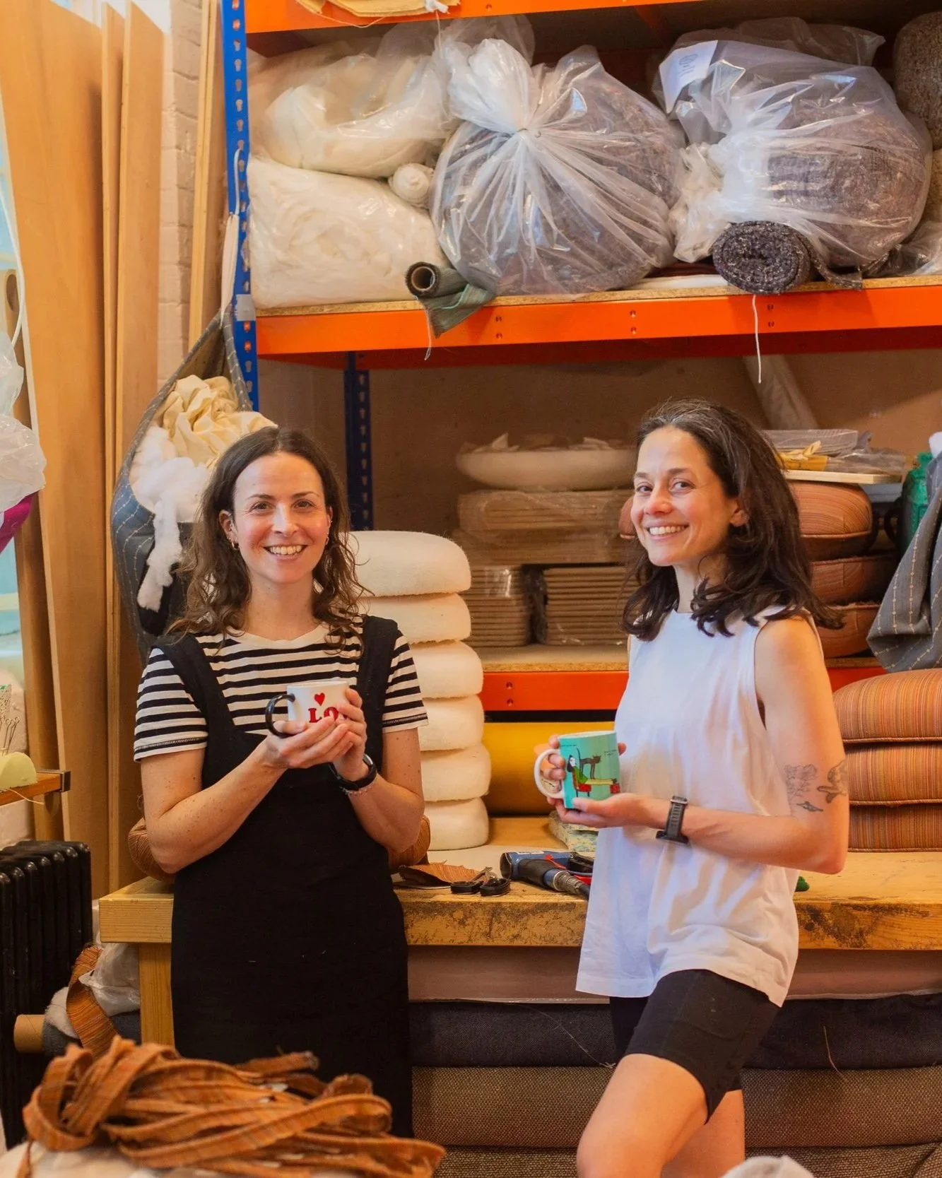 Two women standing and smiling inside a store or workshop, each holding a mug, with shelves of fabric and upholstery materials in the background.