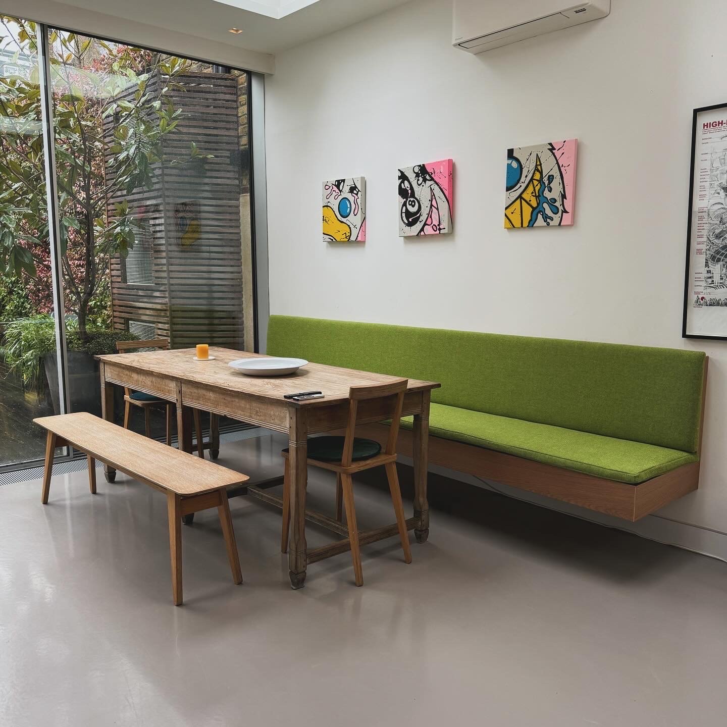 Modern dining area with a wooden table, green upholstered bench, and matching wooden chairs, next to a large window with plants outside, decorated with colorful abstract artwork on the wall.