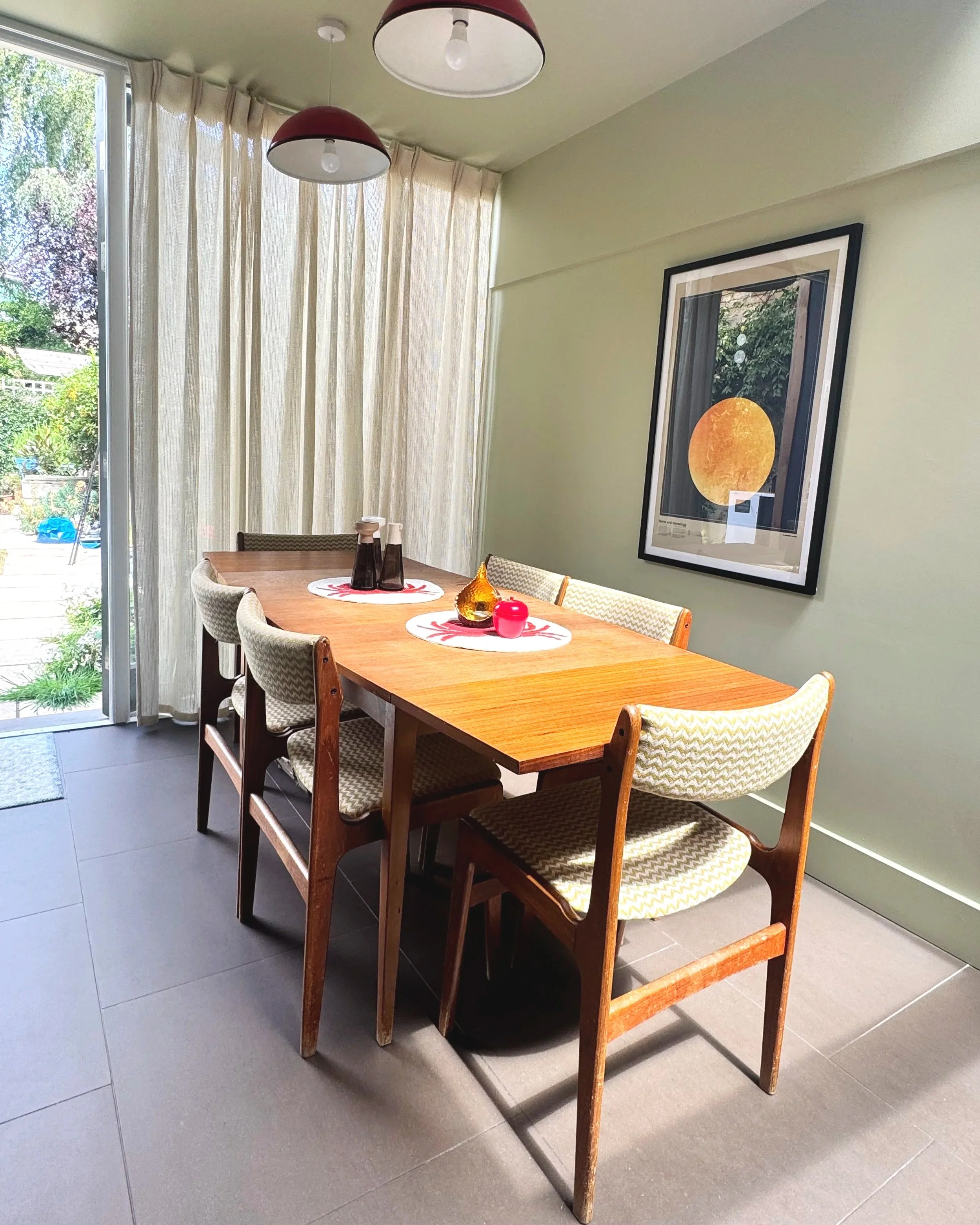 A dining room with a large wooden table surrounded by eight chairs with patterned upholstery. The table has decorative items including a gold-colored vase, a red candle, and black bottles. There are beige curtains over glass sliding doors leading to an outdoor patio. On the wall, there's a framed picture of a large orange moon and trees reflected in a window.