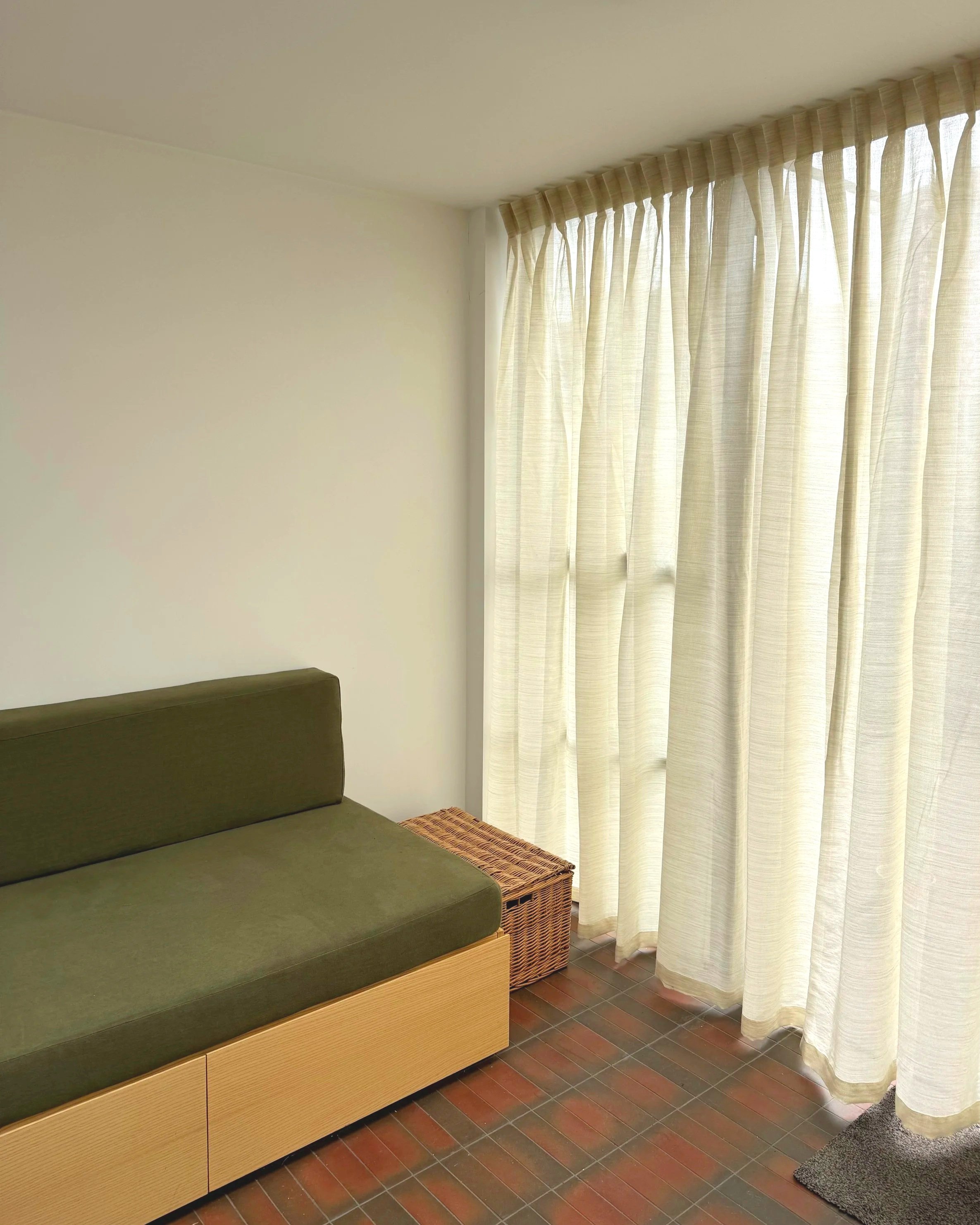 A corner of a room showing a green cushioned bench with a wooden base, a wicker basket under it, white curtains covering a large window, a small gray rug on the wooden floor, and part of a gray carpet.