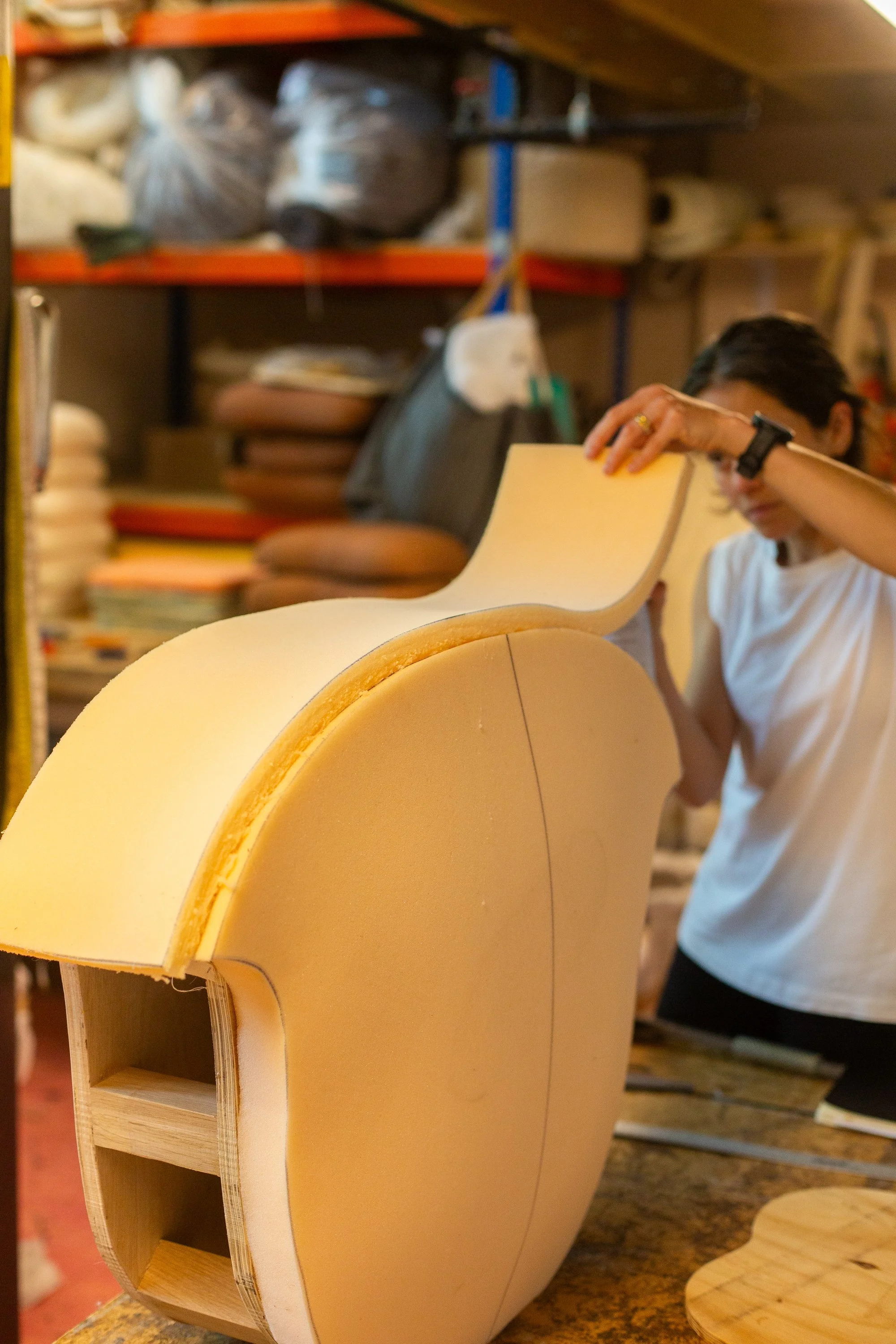 A person working on the upholstery of a wooden seat  in a workshop with shelves of materials in the background.
