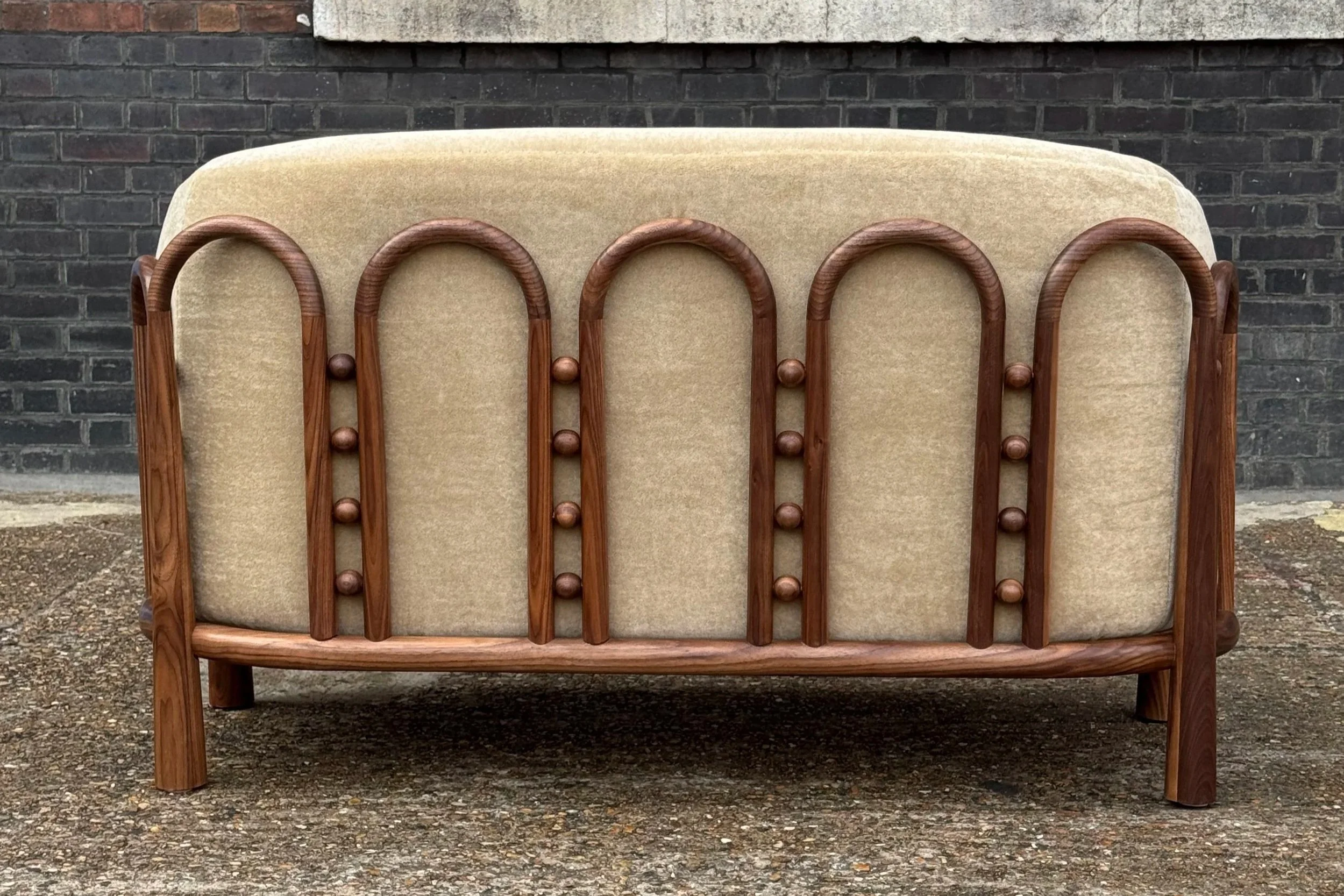 A handmade walnut sofa, with curved and turned exposed wood frame with a beige upholstered backrest and seat, situated outdoors on a rough concrete surface in front of a dark brick wall with a concrete ledge above.