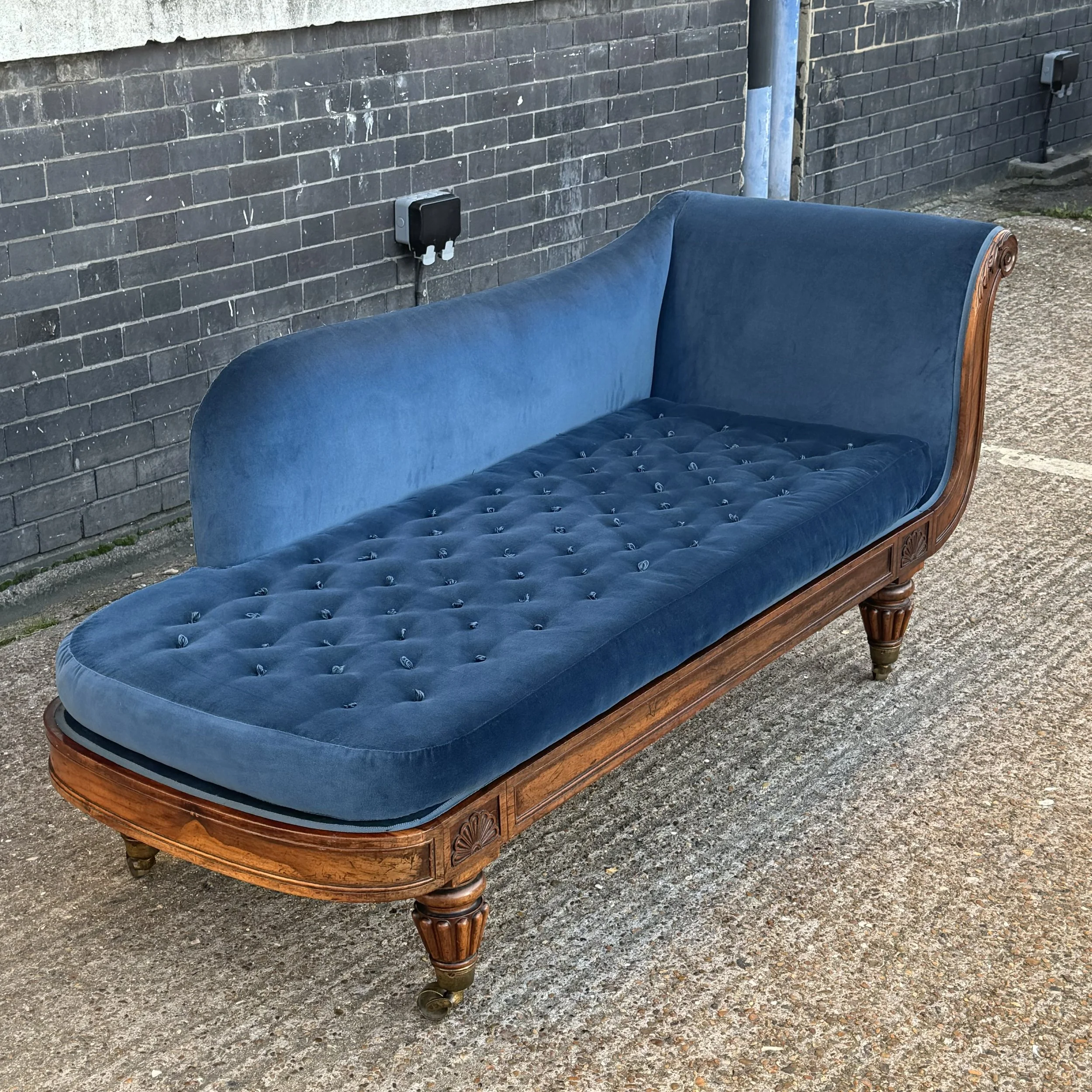 A vintage wooden sofa with blue upholstery and carved wooden accents, placed outdoors on a gravel surface against a dark brick wall.