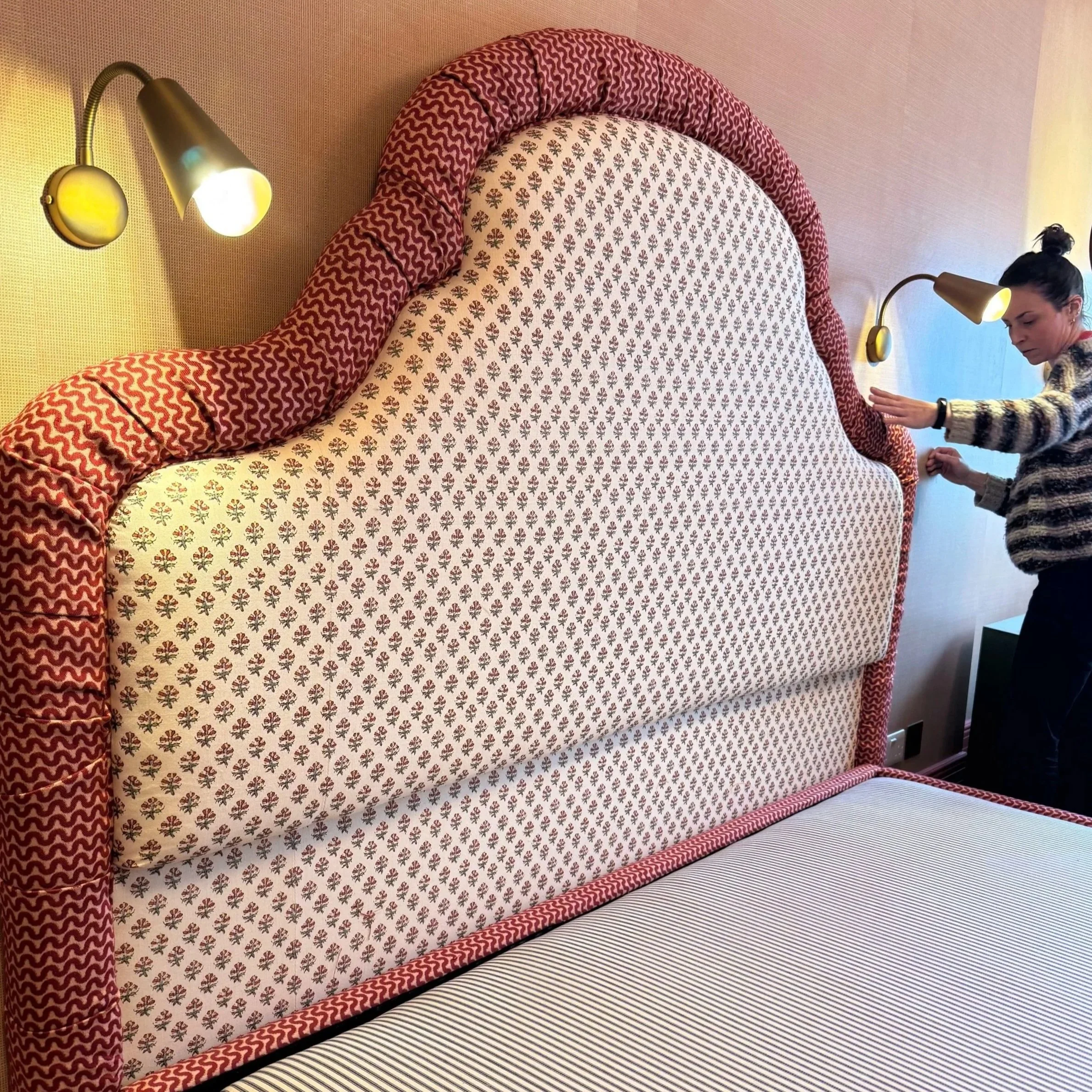 Close-up of a bed with a decorative headboard, red and white patterned upholstery, and a striped bedsheet. A woman is adjusting the headboard, standing beside it.