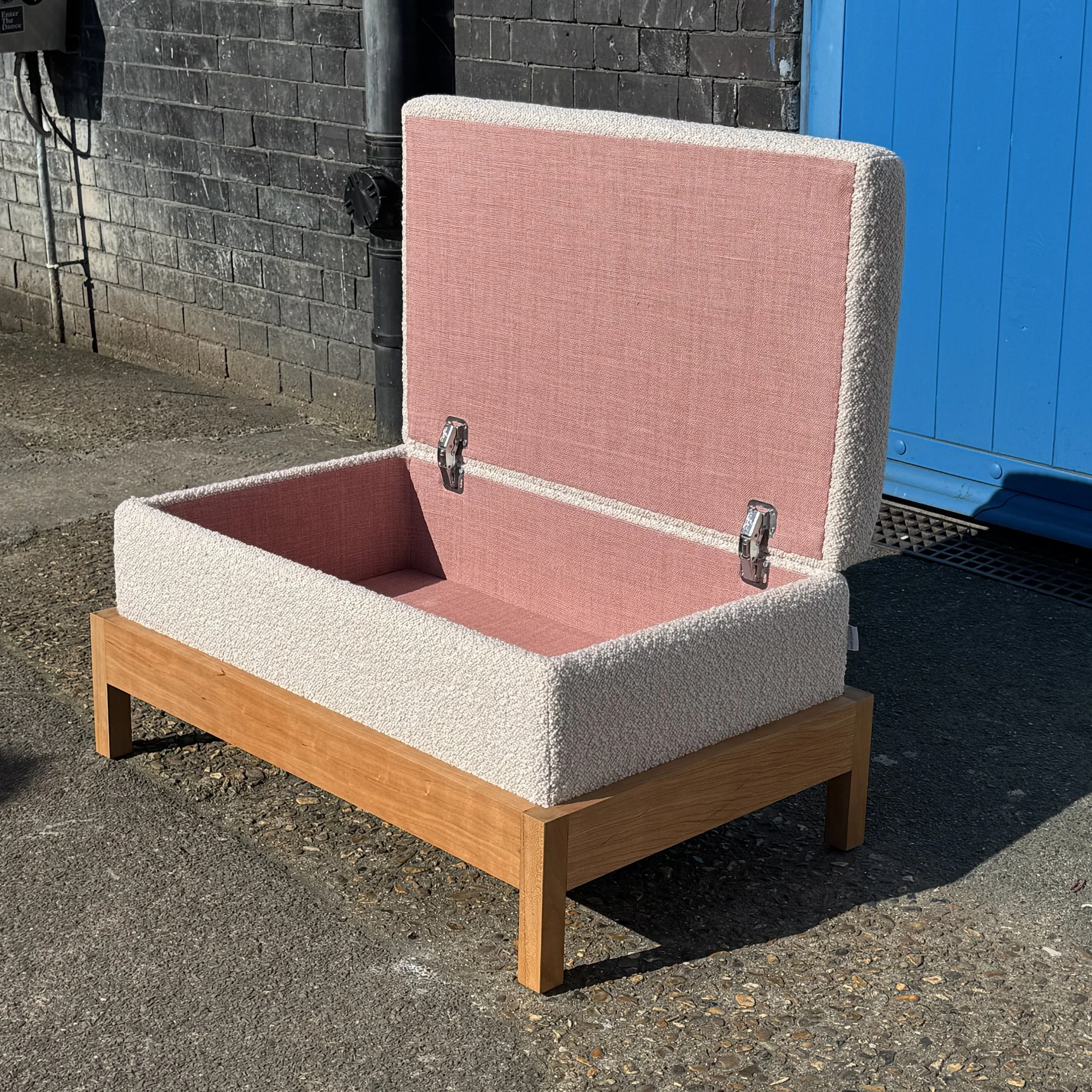 An open box ottoman with cherry wood base upholstered in a white boucle fabric with a pink interior against a brick wall and blue door.