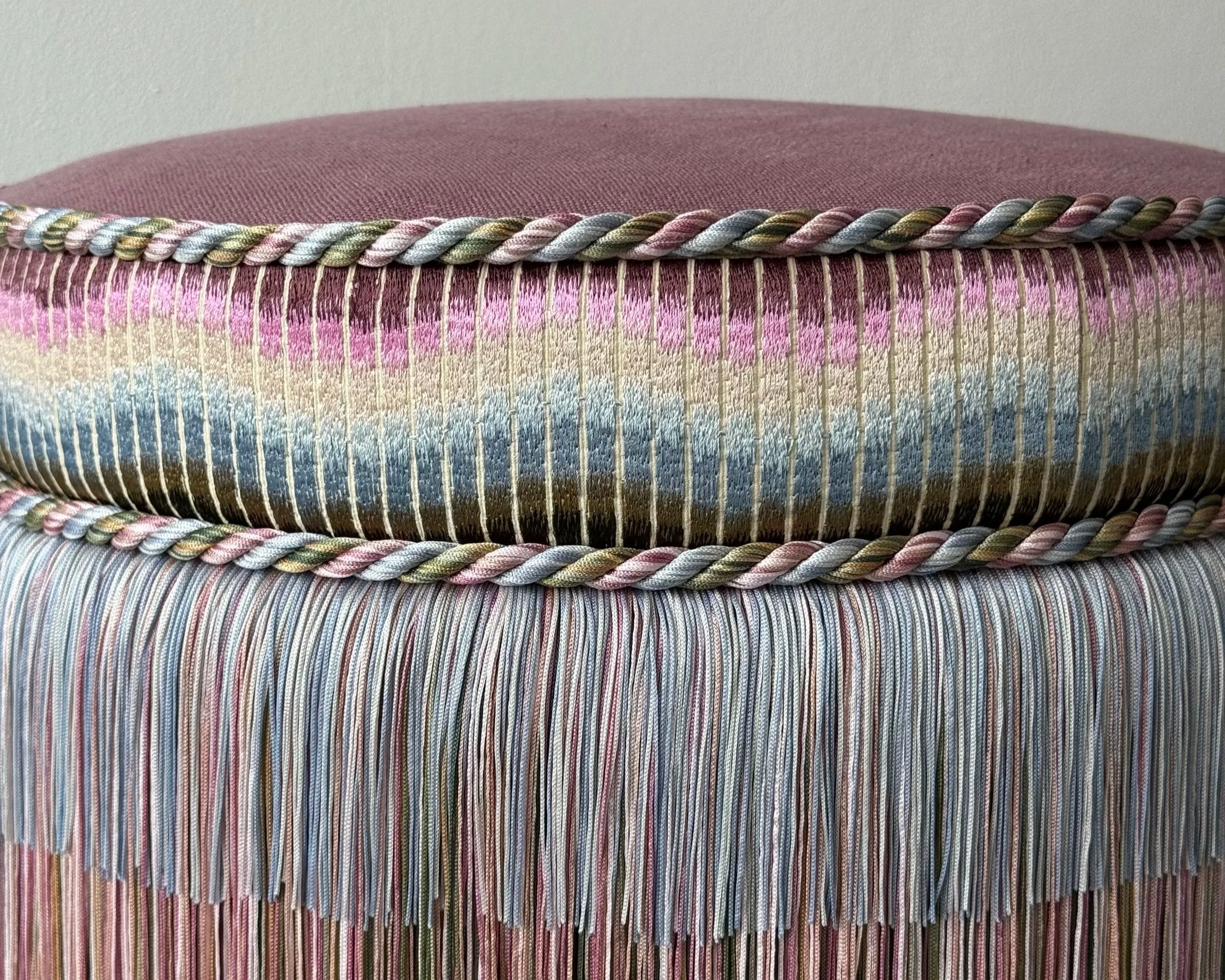 Close-up of a woven textured fabric with horizontal multicolored stripes in shades of pink, purple, beige, blue, and green, bordered by a braided trim on top.