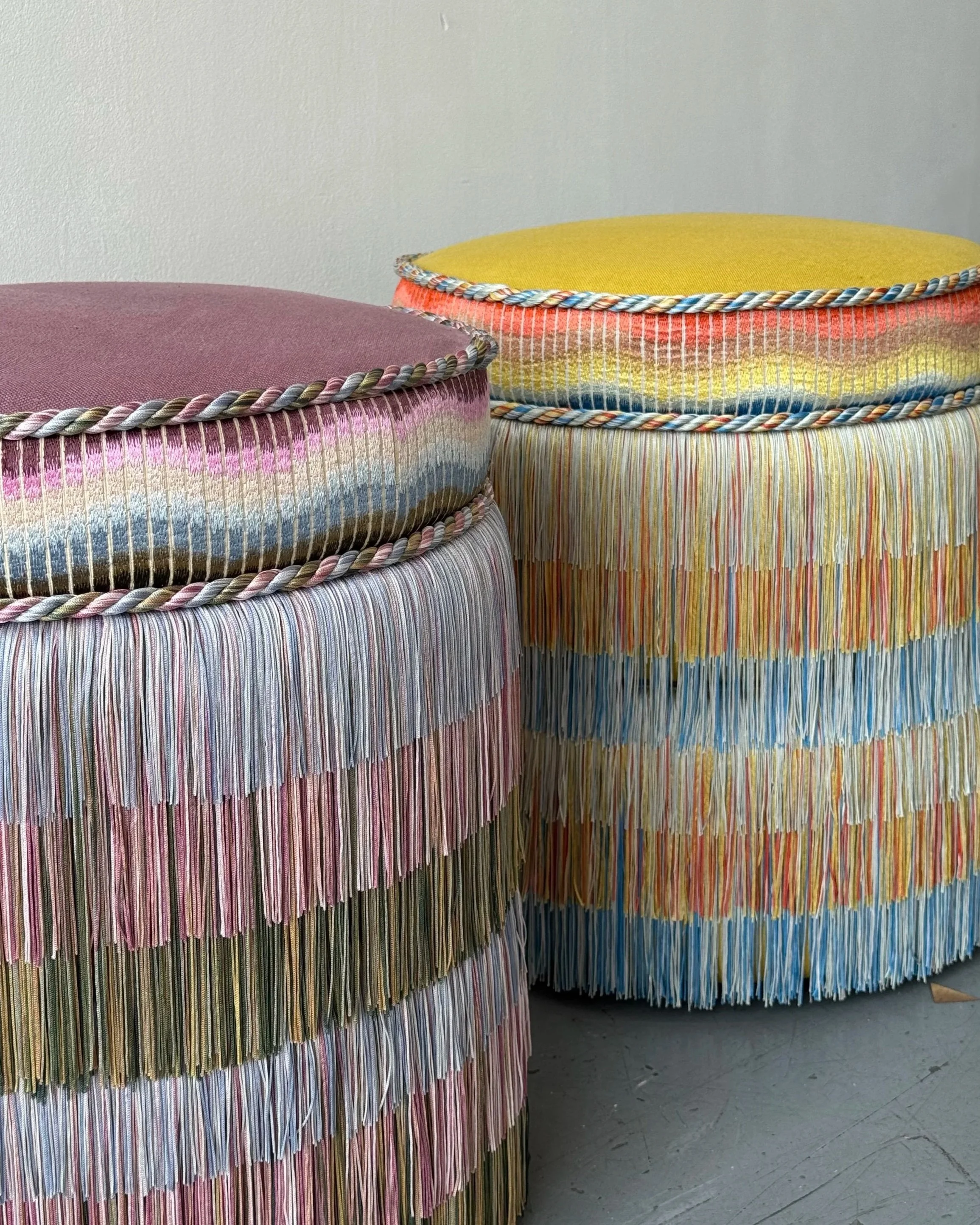 Two large cylindrical sculptures made from colorful, vertically hanging paper strips with fringed edges, topped with rounded fabric cushions, in an art installation against a plain wall.