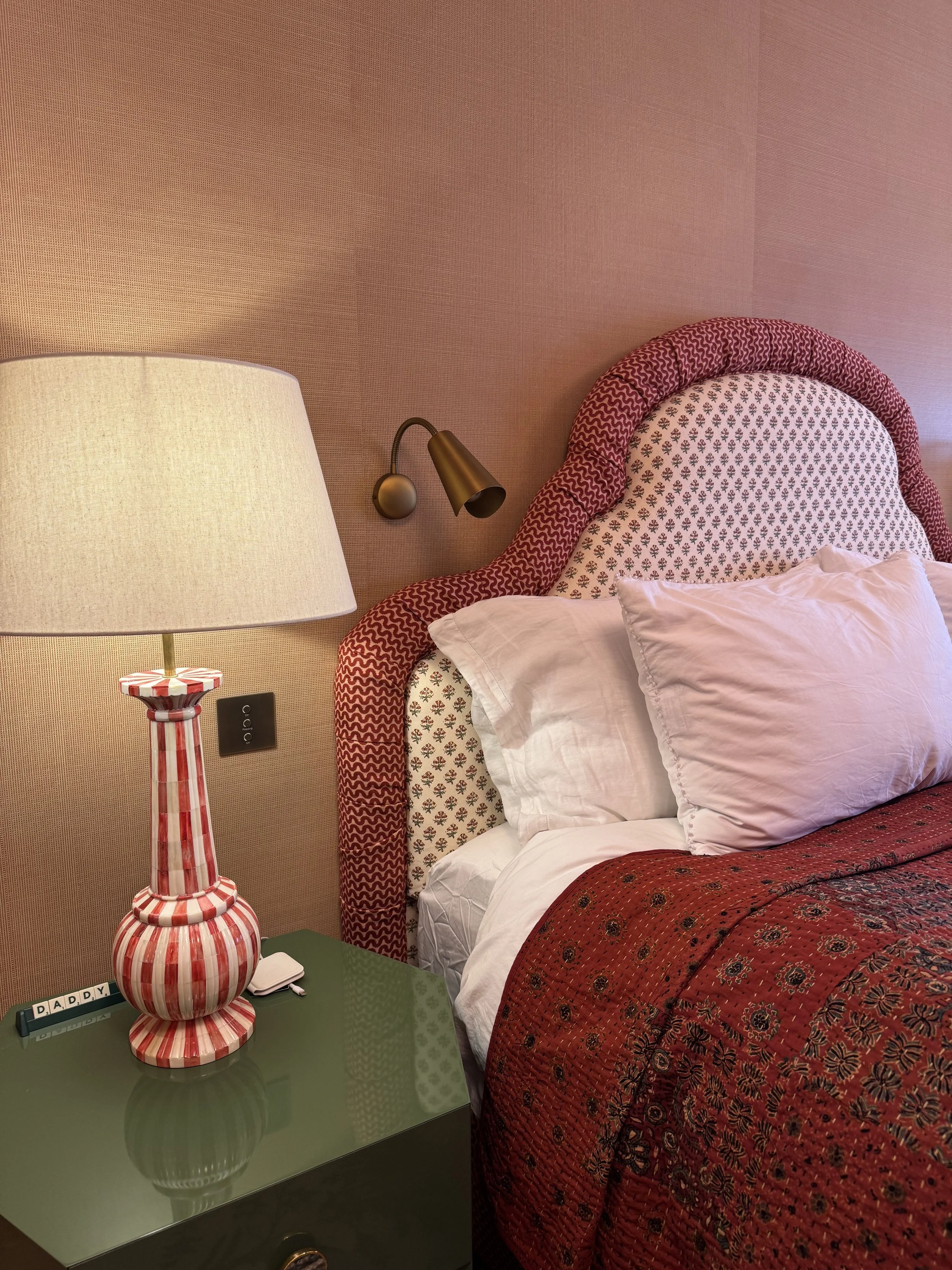 A bedroom with a patterned pink headboard, white pillows, a red quilt, a green side table, a large white lampshade lamp, and a small black alarm clock.