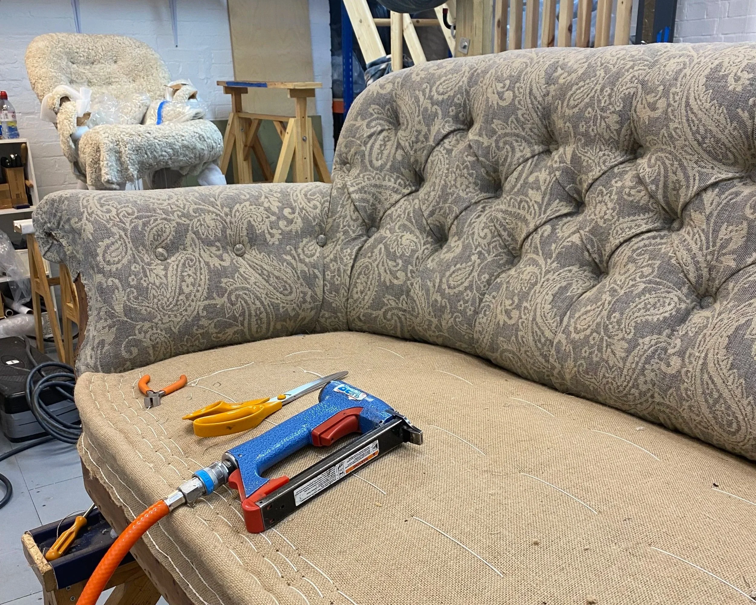 A workshop scene with a beige and brown patterned sofa, a disassembled armchair wrapped in plastic, a wooden small table, and various tools including a saw, pliers, and a drill on the sofa's seat. There are also cords and other miscellaneous tools and objects around the workspace.