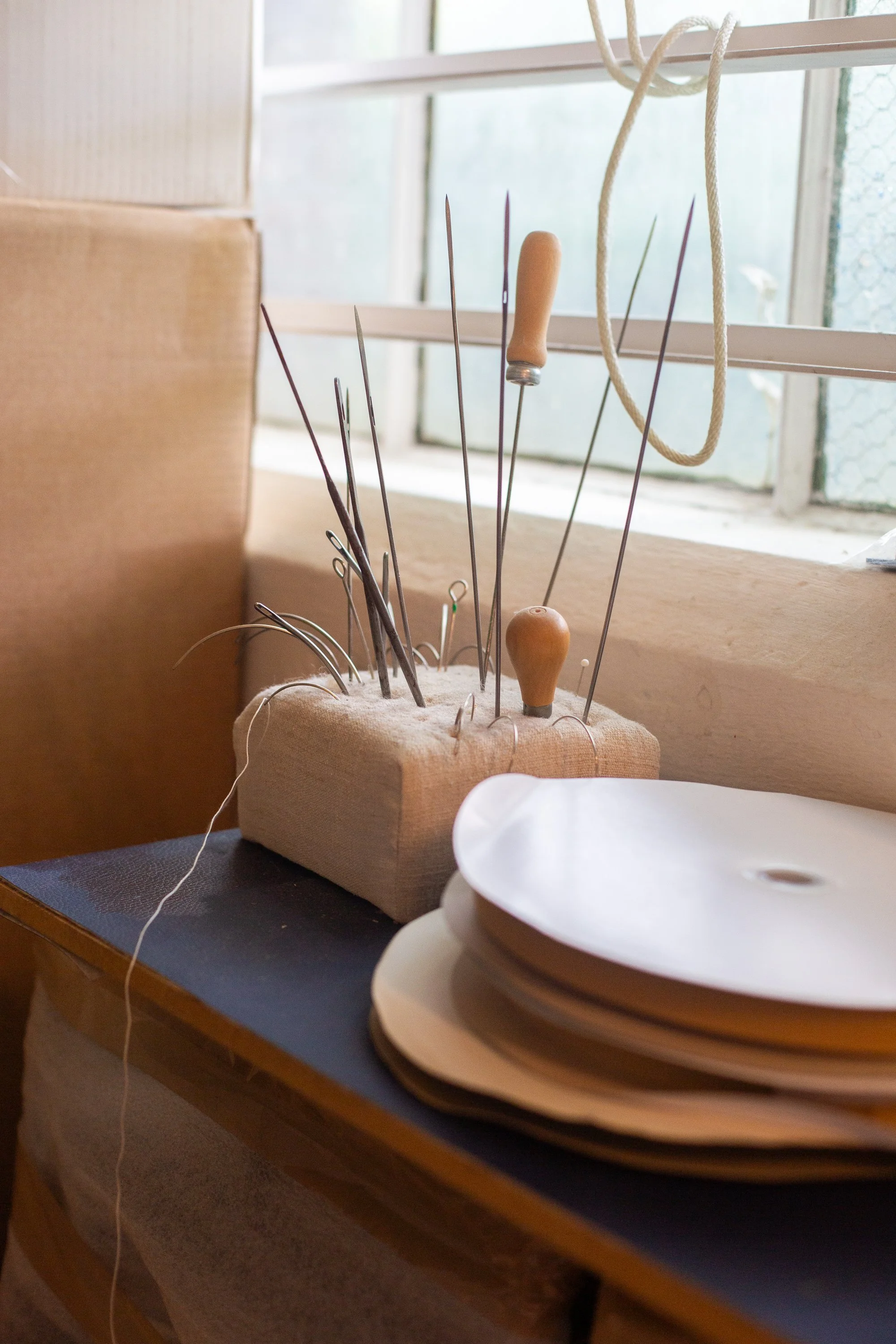 A collection of sewing or upholstering tools with wooden handles, metal tips, and some with looped wire, arranged in a foam holder, next to a stack of white spools of thread, near a window with a wire mesh screen.