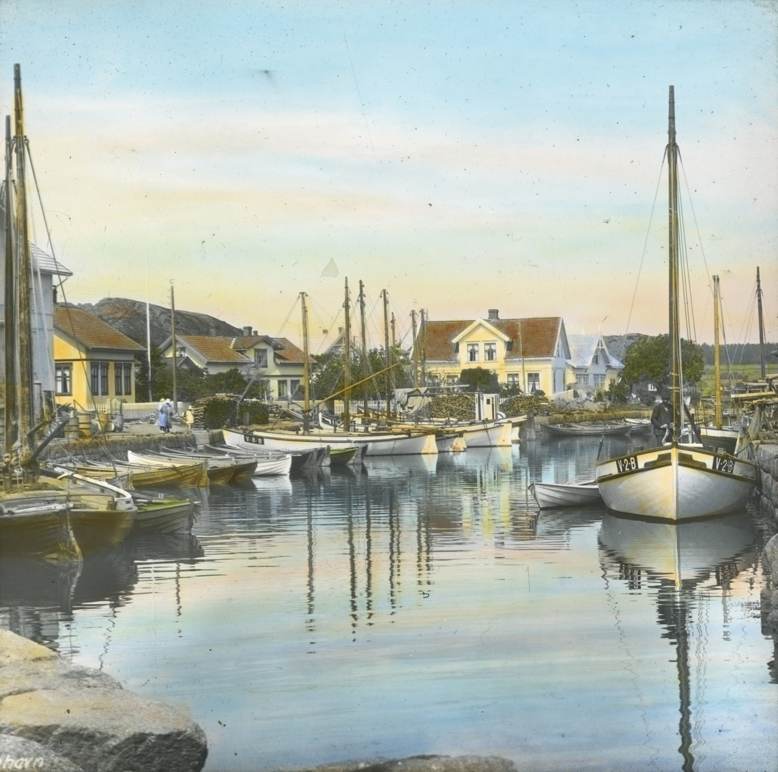  Håndkolorert dias. Seilbåter og robåter ligger fortøyd til kai i Nevlunghavn. I bakgrunnen sees hvitmalte trehus.  Fotograf: Wilse, Anders Beer / Norsk Teknisk Museum 