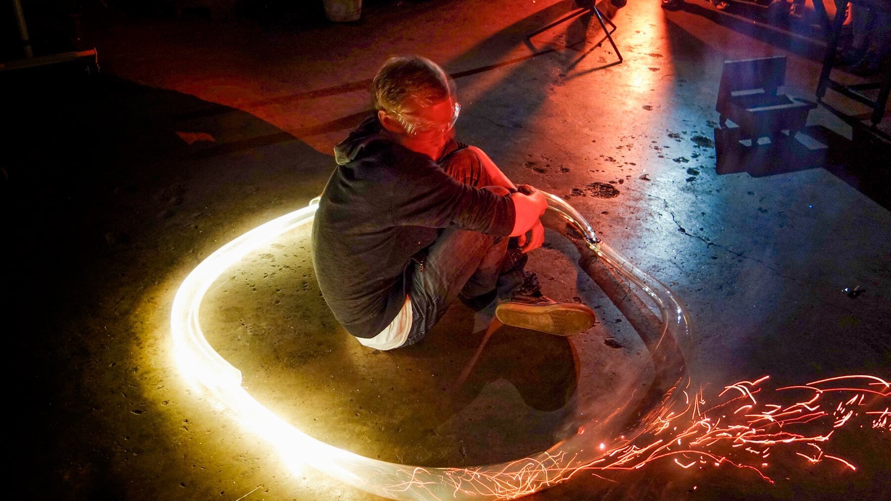 A man sits inside a ring of glowing glass and sparks