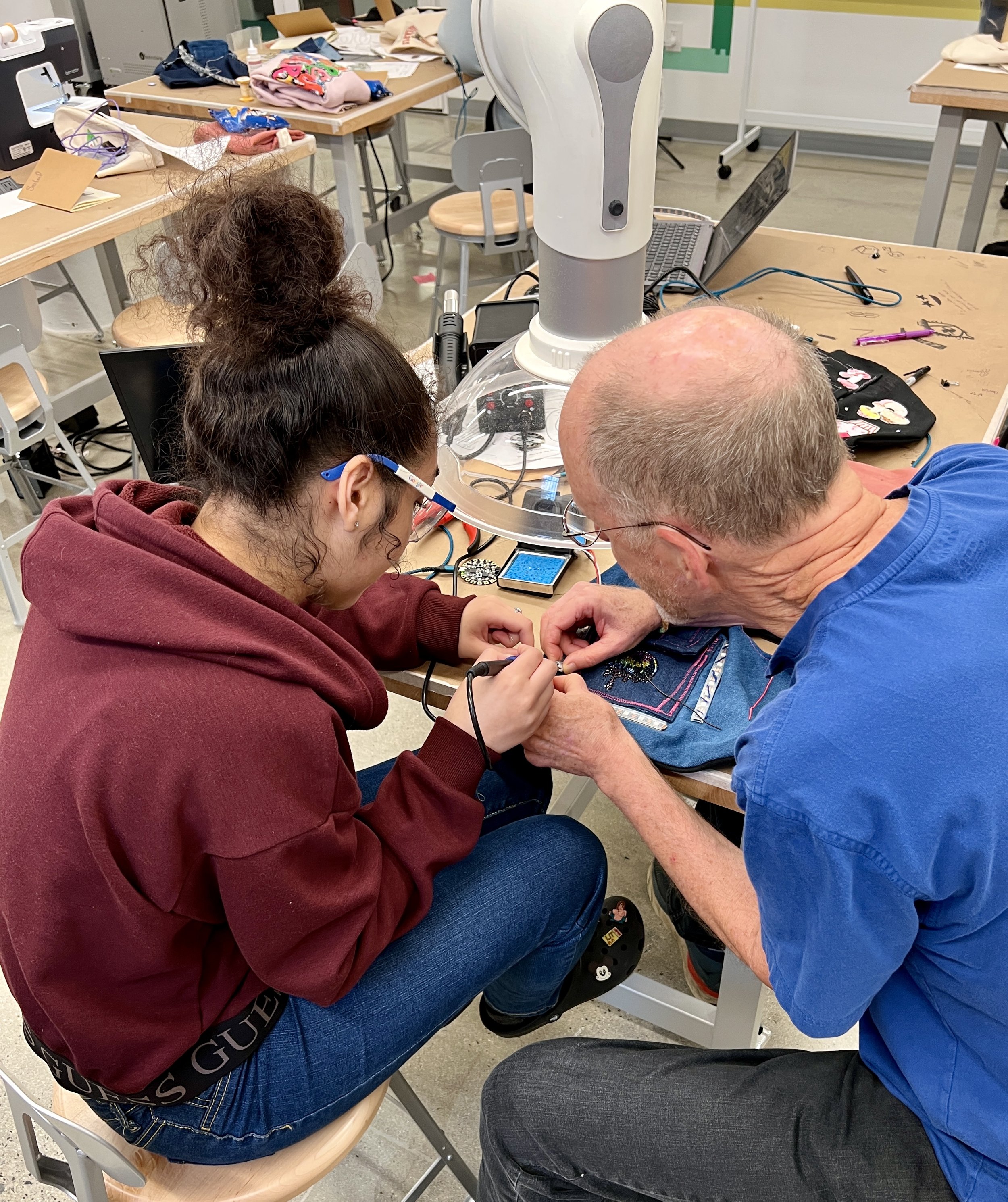  Rob Hart, in the blue shirt, leans in to help a student with an e-textiles project at the lab's workbench, surrounded by tools and materials for crafting and innovation. 