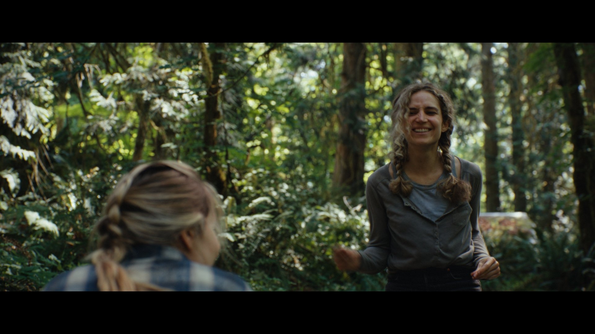 Two women with braided hair are in a forest, one is sitting with her back to the camera, and the other is standing and smiling.