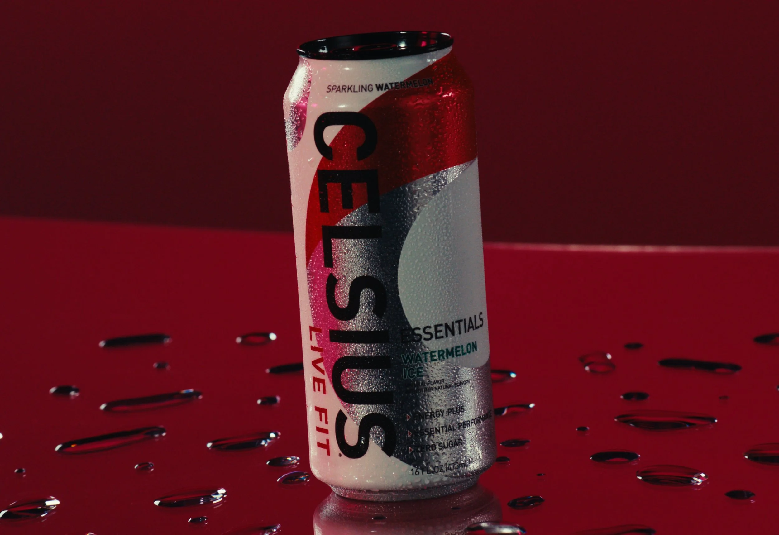 A can of Celsius Sparkling Watermelon flavored energy drink with condensation on the surface, placed on a reflective red surface with droplets of water around it, against a red and dark background.