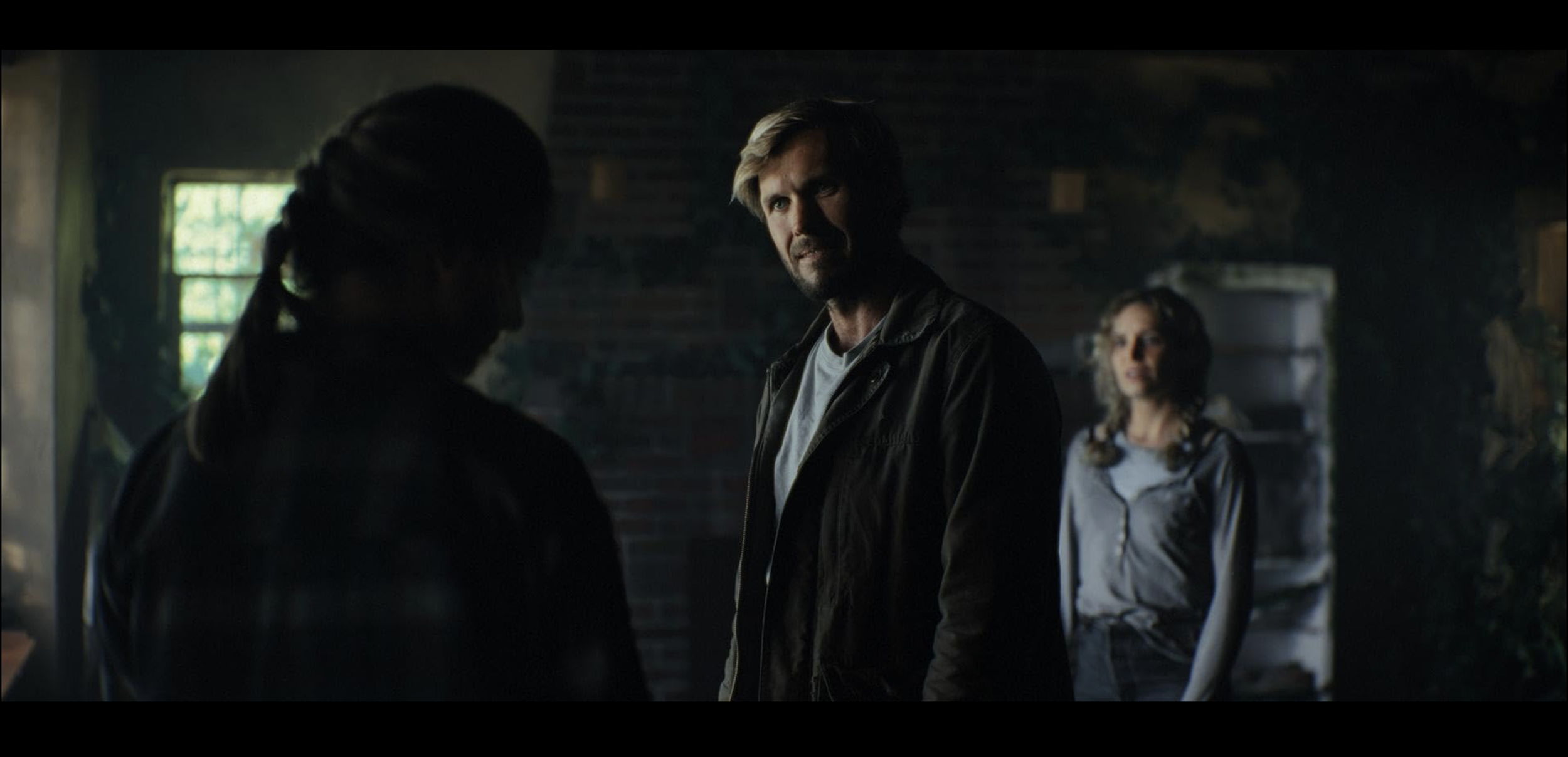 Three people having a conversation in a dimly lit room with a window in the background. The man in the center has blond hair and a beard, wearing a black jacket. The woman on the right has curly hair and glasses, wearing a gray top. The woman on the 