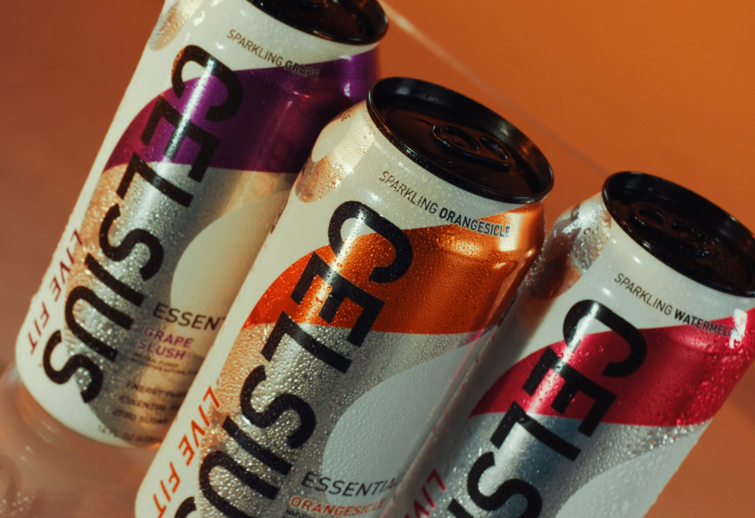 Three cans of Sparkling Water with condensation on the cans. The cans are arranged diagonally, with one purple grape flavor, one orange citrus flavor, and one watermelon flavor, against an orange background.