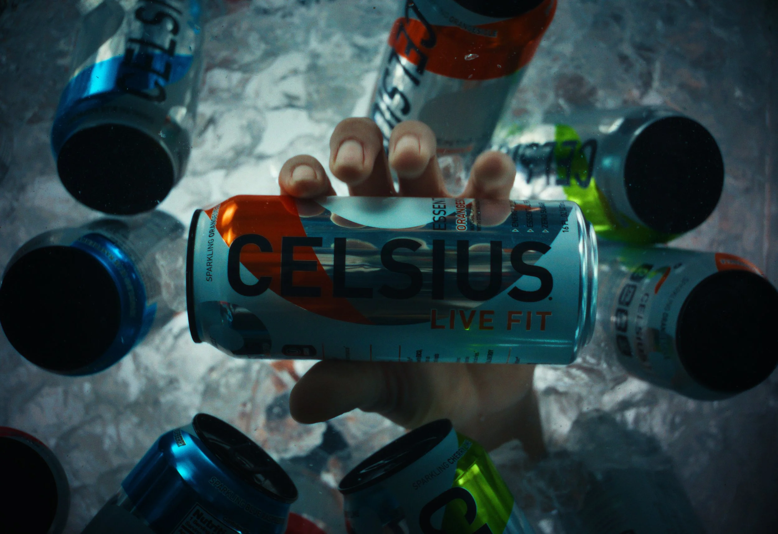A hand holding a can of Celsius energy drink amid a scattered arrangement of other Celsius cans on a textured surface.