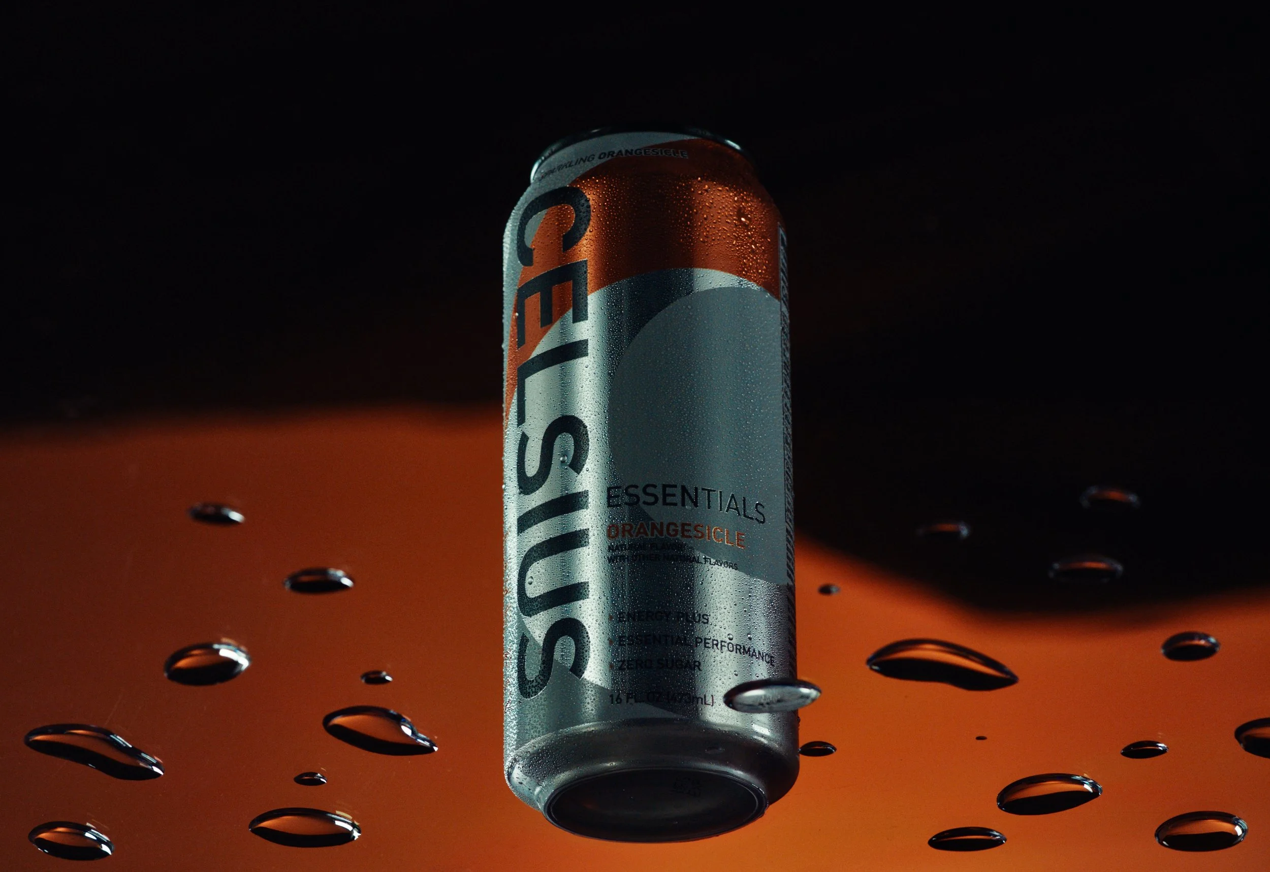 A can of Celsius Essentials Orange Sicle flavored energy drink with droplets of condensation on the can, floating above a mostly orange gradient background with scattered water droplets.