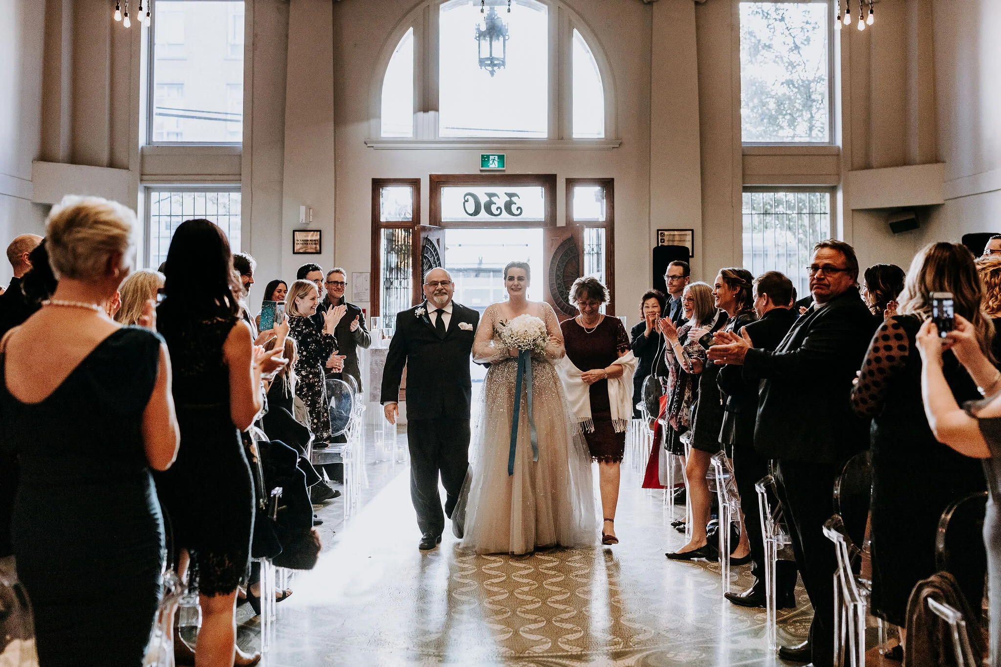 Bride walking down the aisle with two companions, surrounded by guests in a well-lit wedding venue, guests applauding.