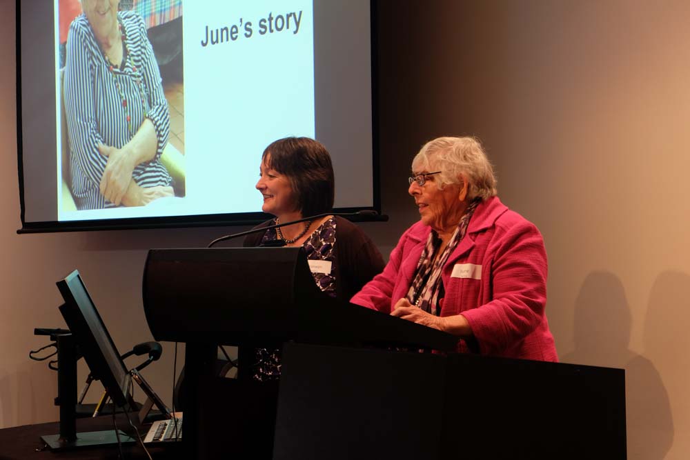  Two older women are standing behind a lectern and presenting at a conference.  A projector screen can be seen in the background with a photo of one of the women with the words ‘June’s Story’. 