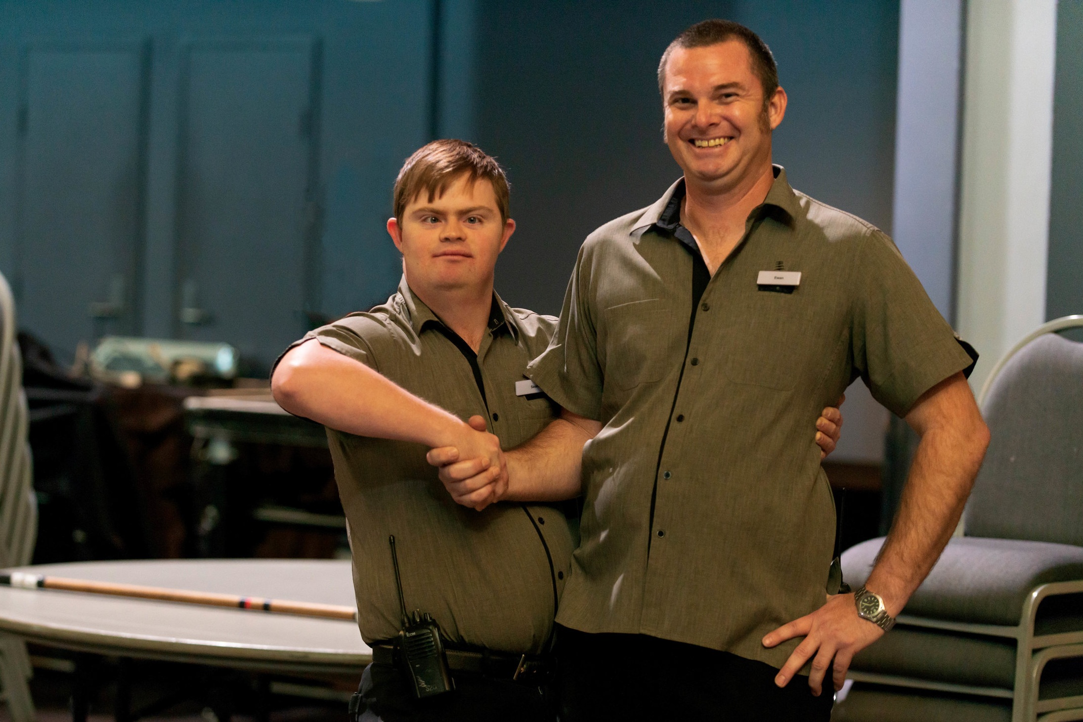  A man with disability and his colleague shaking hands and smiling.  They wearing uniforms for the Brisbane Convention center and surrounded by the equipment they need for work. 