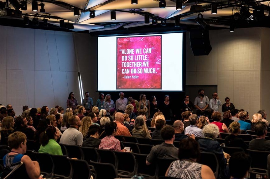 A photo from a conference.  It shows a large group of people watching a presentation.  There’s a slide projected on the wall that reads ‘Alone we can do so little, together we can do so much’. 