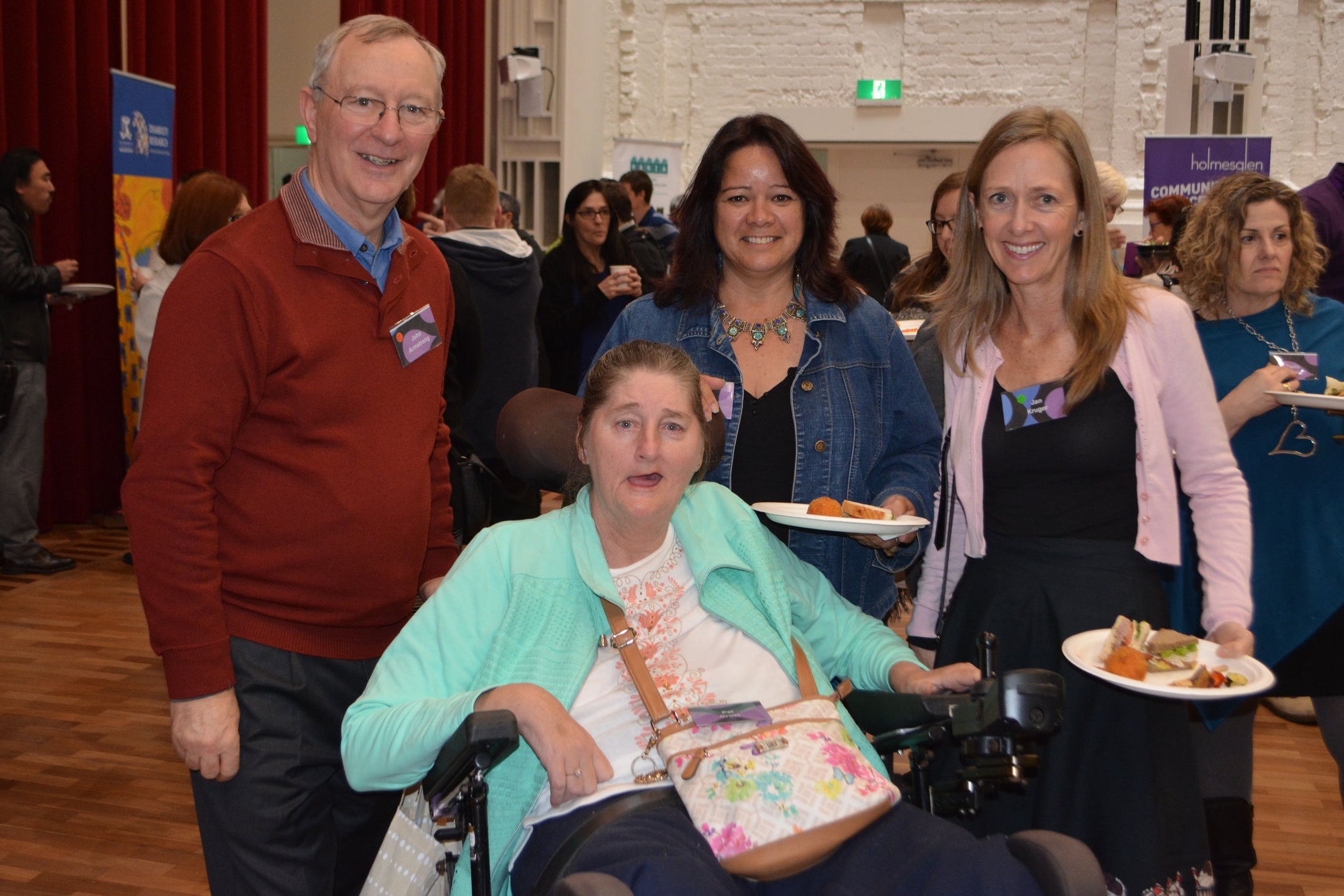  Four attendees at a conference standing with their lunch in hand smiling at the camera.  There’s a man and three women.  One of the women is in a wheelchair. 