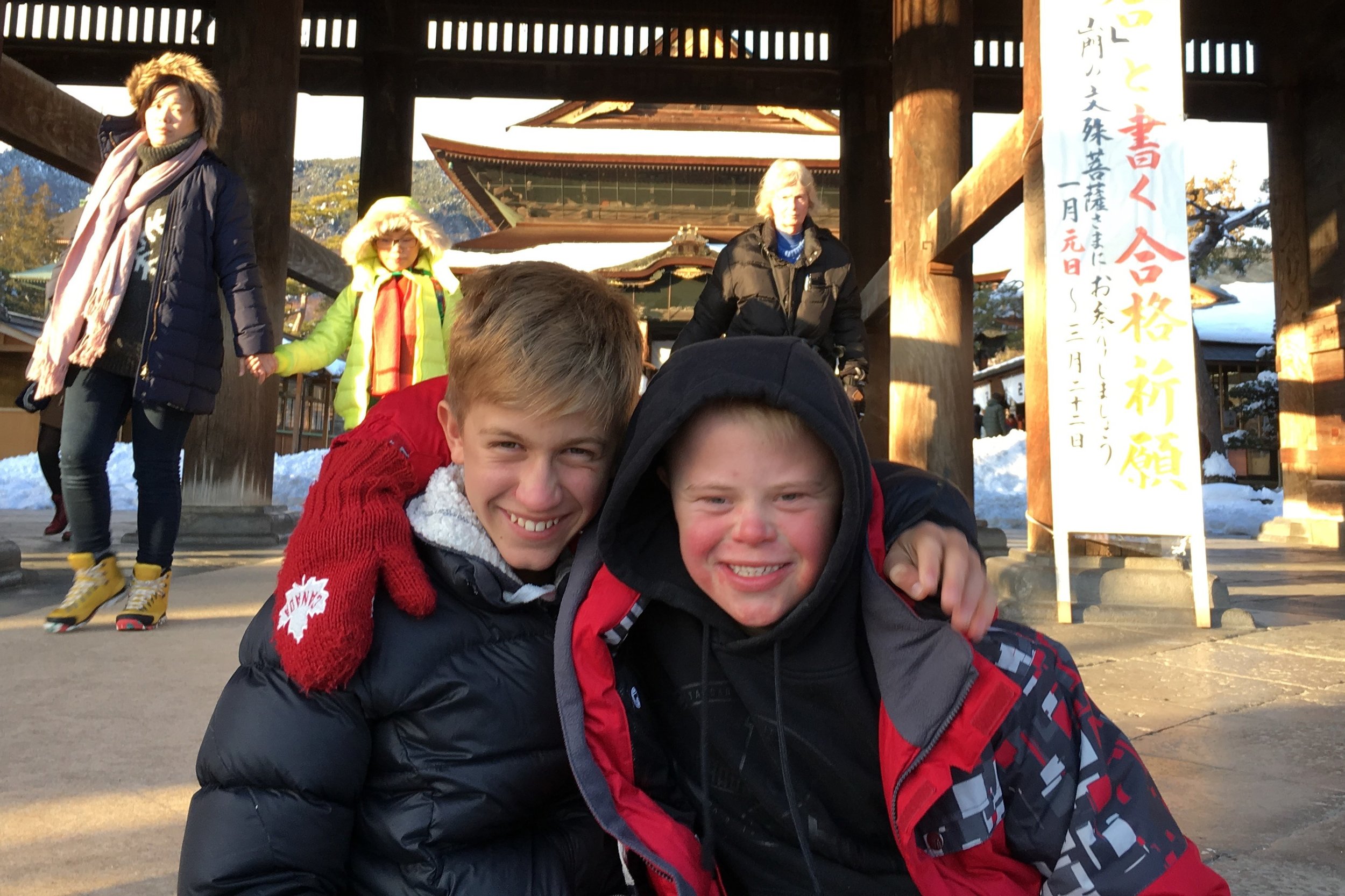  Two young men are facing the camera and wearing thick jackets and gloves, smiling and hugging in front of a temple in Japan.  There’s Japanese writing behind them.  The boy on the right has a disability. 